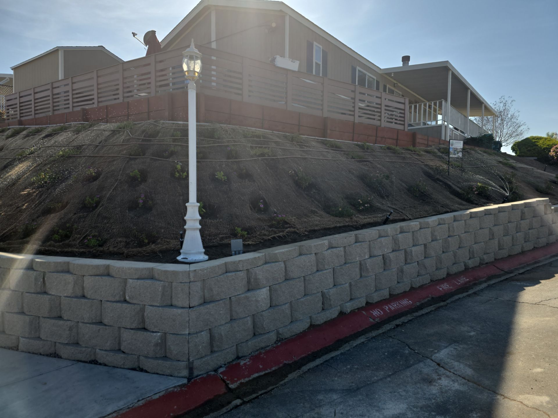 Retaining wall of grey blocks supports hillside with house, street curb with red line, and decorative lamp post.
