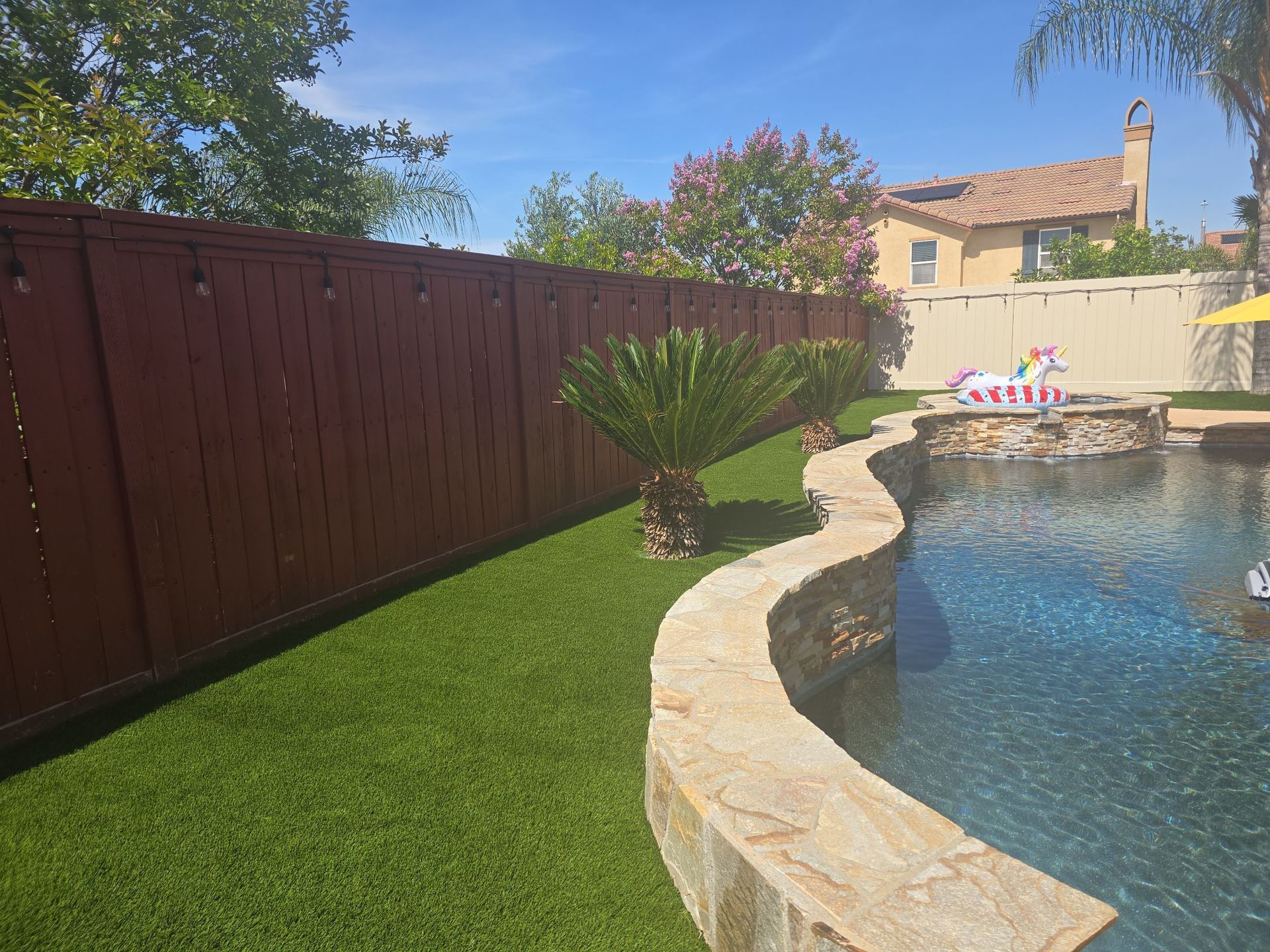 Backyard with a brown wooden fence, green lawn, pool, and a sunny sky.