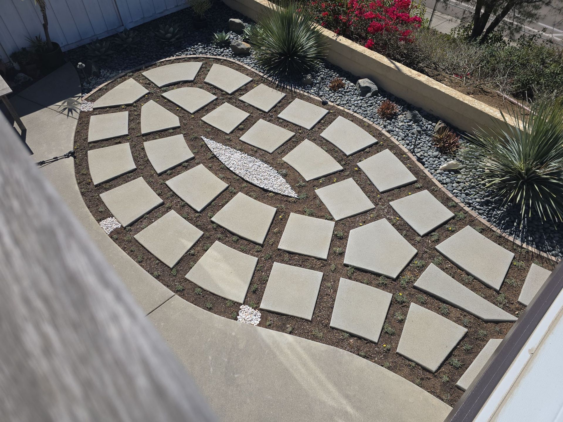 Overhead view of a patio with a stone pattern and gravel border, surrounded by vegetation and concrete.