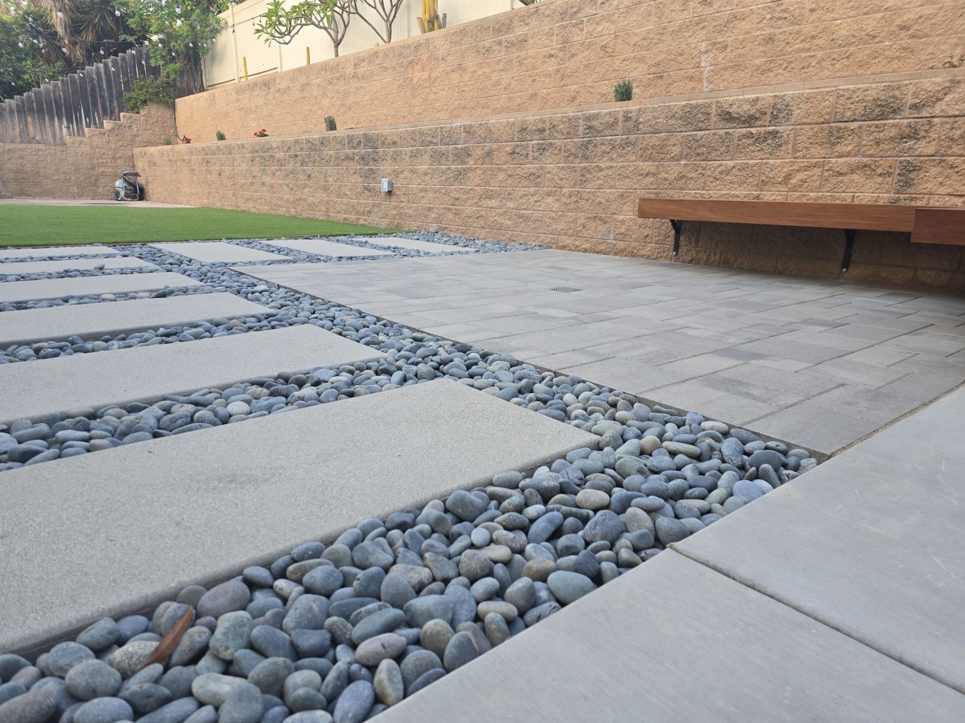 Stone pavers with grey river rocks, retaining wall with a bench and grass.