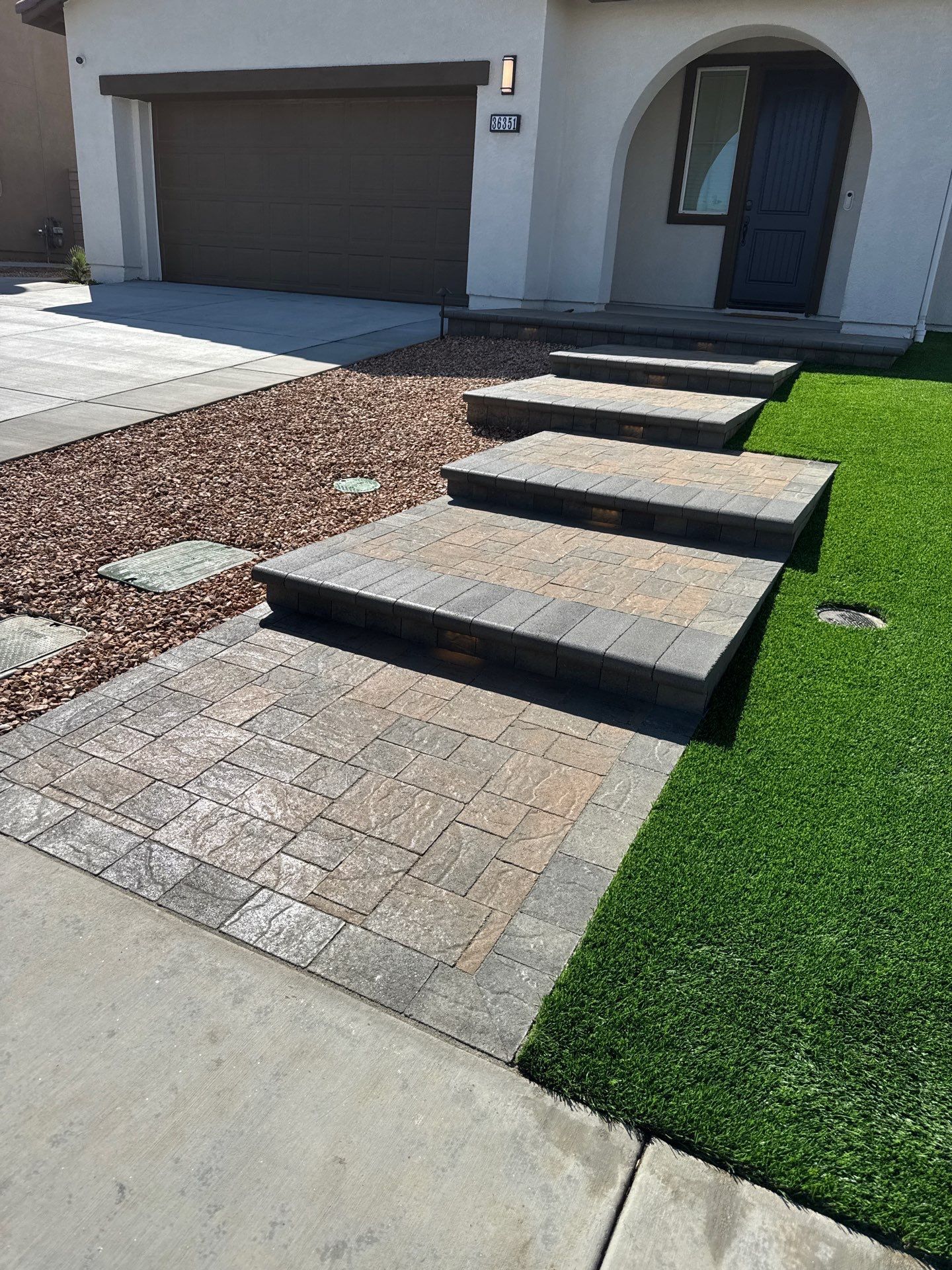 Brick walkway with steps leading to a house entrance. Green grass and gravel border the pathway.