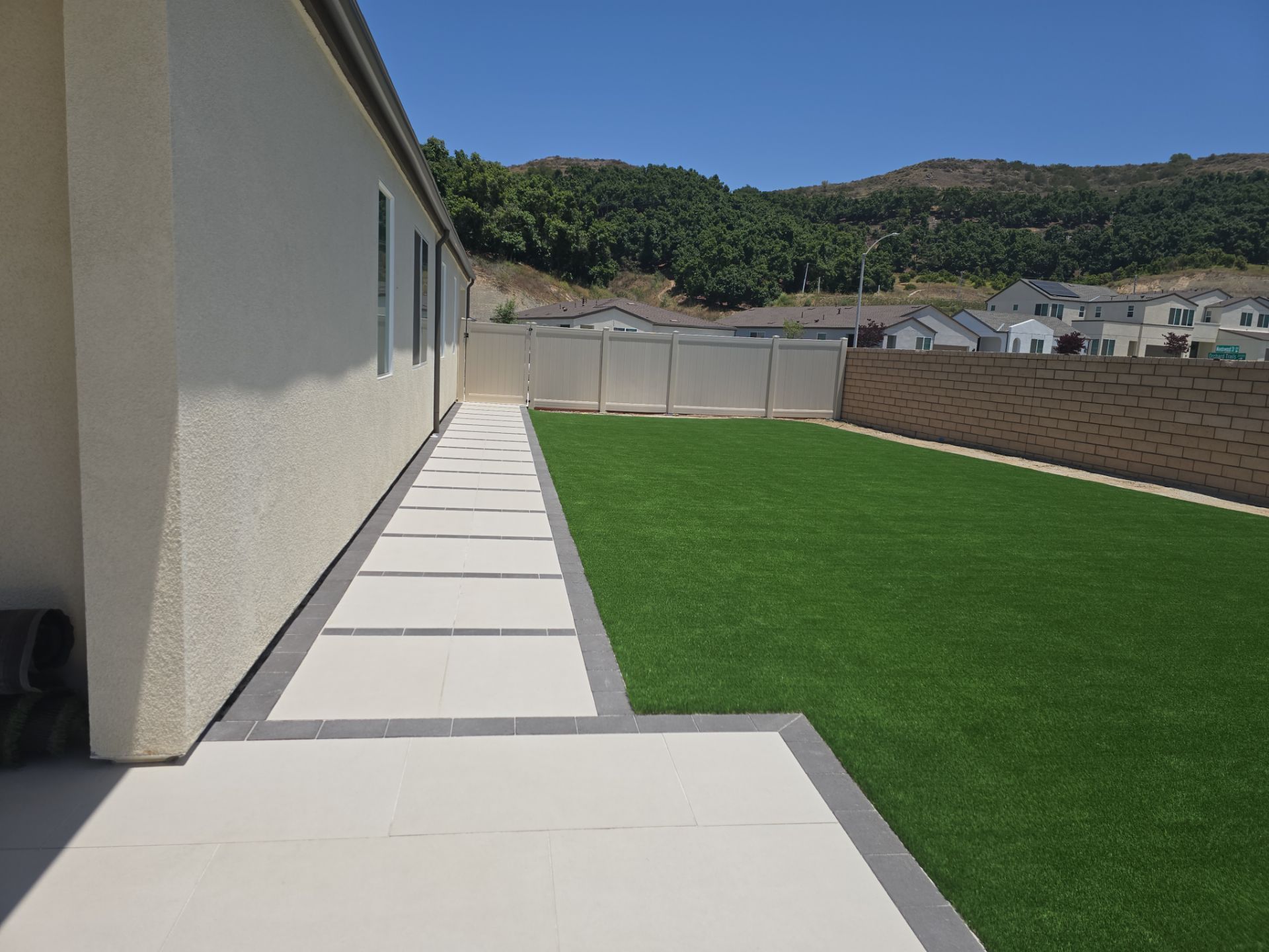 Backyard with faux grass, concrete walkway along house, fence, and hillside.