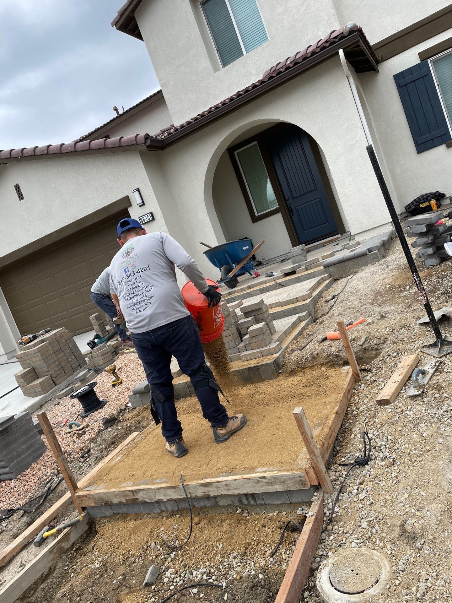 A construction worker compacts gravel with a machine, preparing a pathway in front of a house.