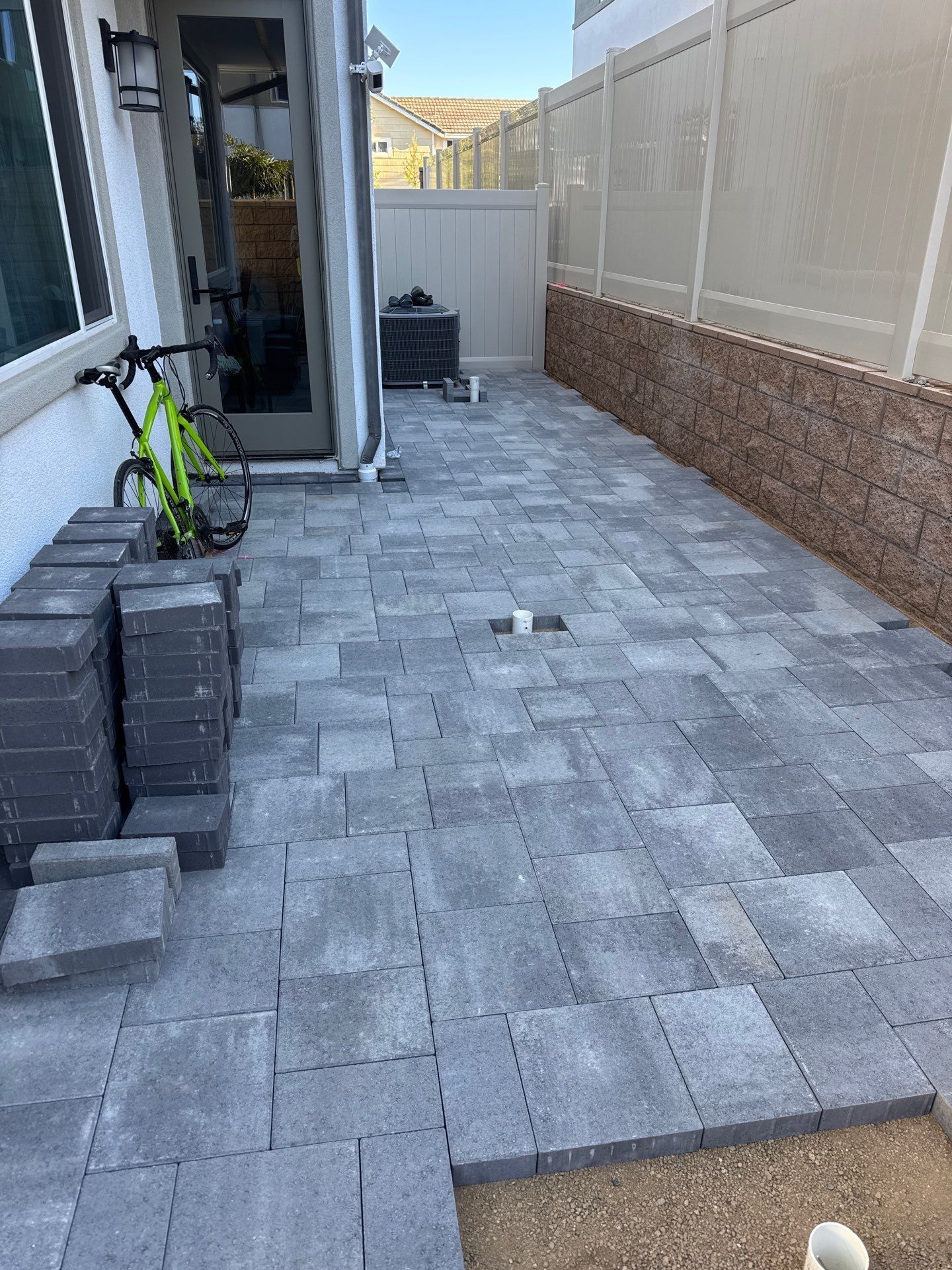 Patio paving in progress with gray bricks. Green bicycle by door. Brown and white wall.