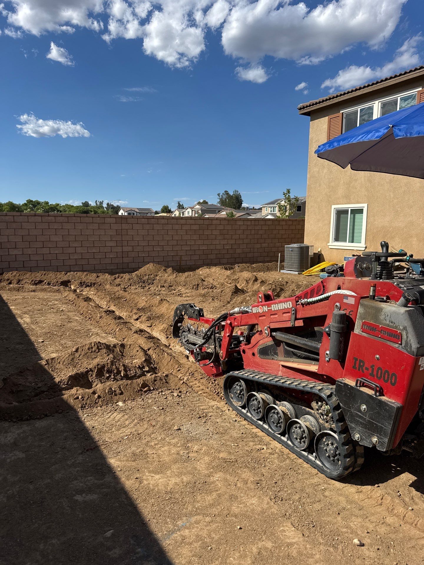 Red compact loader digging soil in a backyard beside a tan house and cinderblock wall under a blue sky
