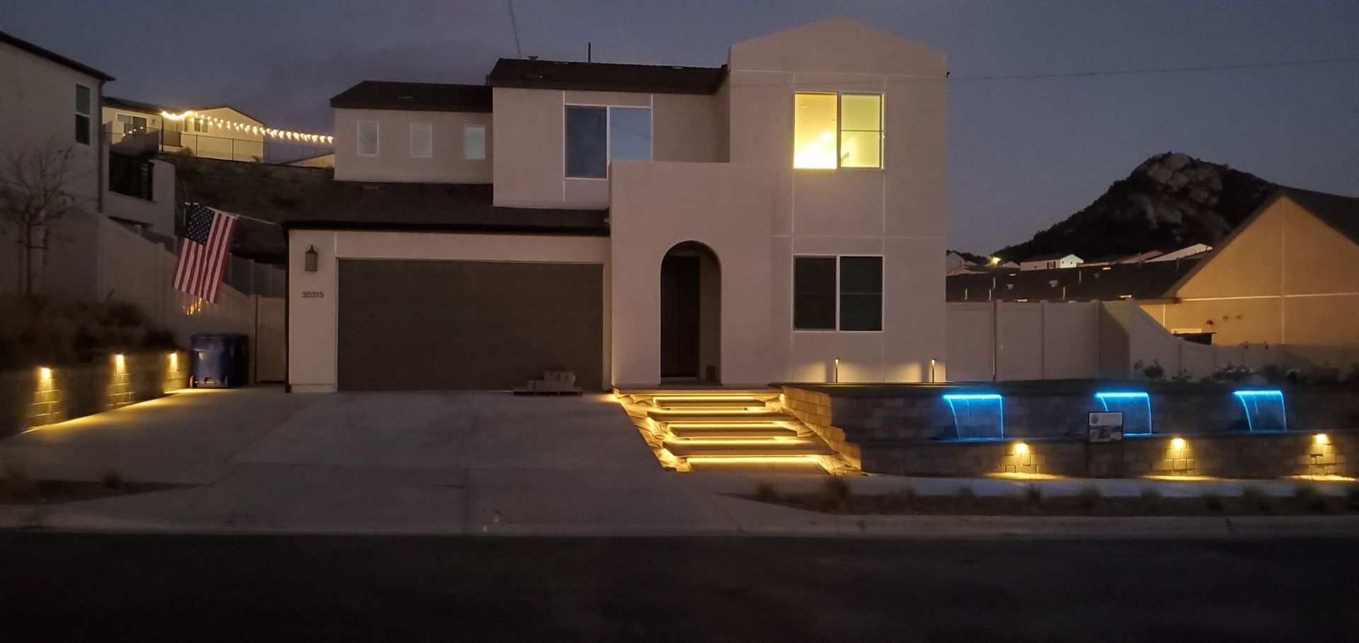 A two-story house at dusk, with exterior lights illuminating steps and landscaping. An American flag is visible.