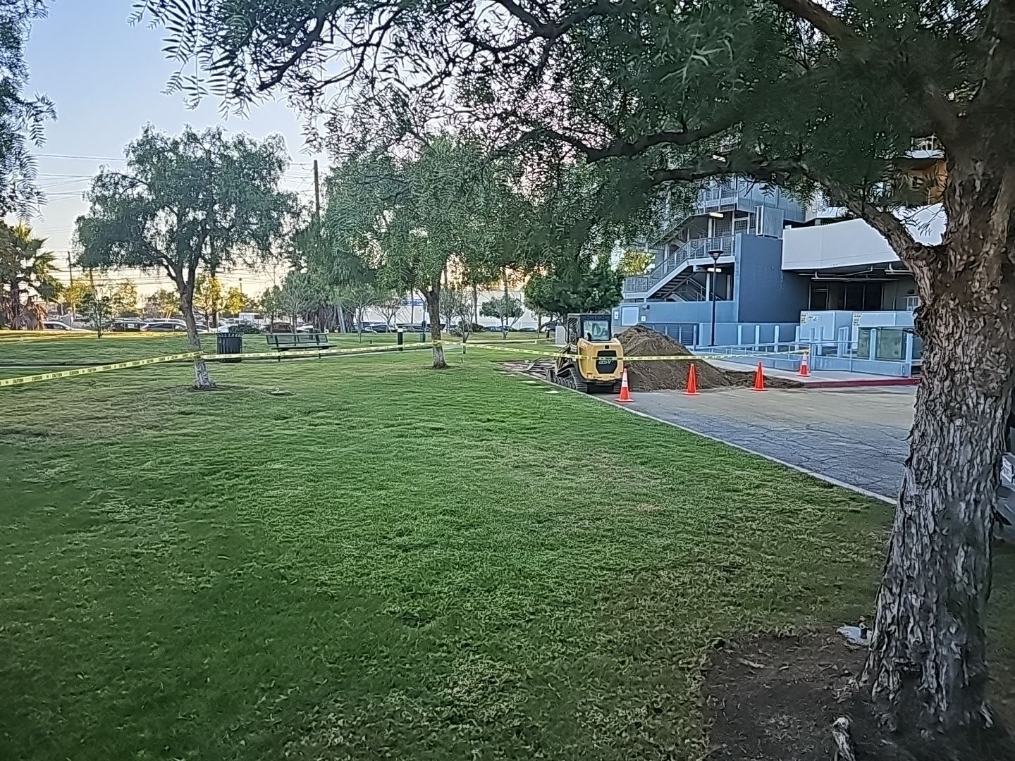 Green park with construction near a light blue building, trees, and an excavator.