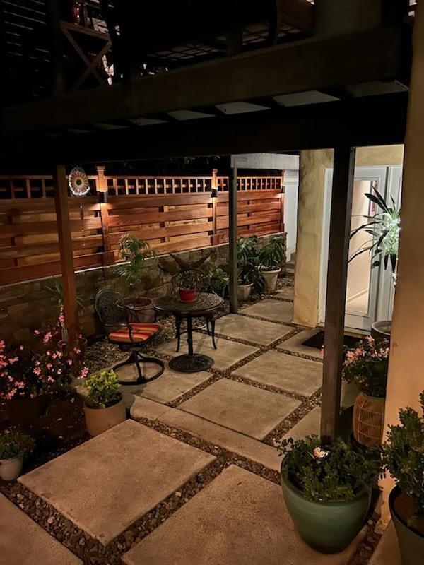 Lit backyard patio with seating, potted plants, wood fence, and pergola.