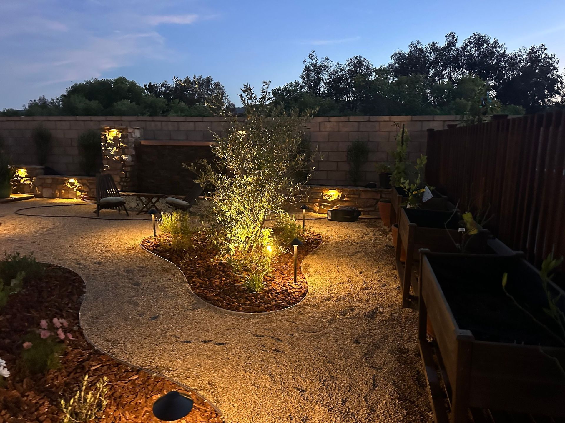Lit backyard garden with gravel path, glowing landscape lights, and raised planters at dusk