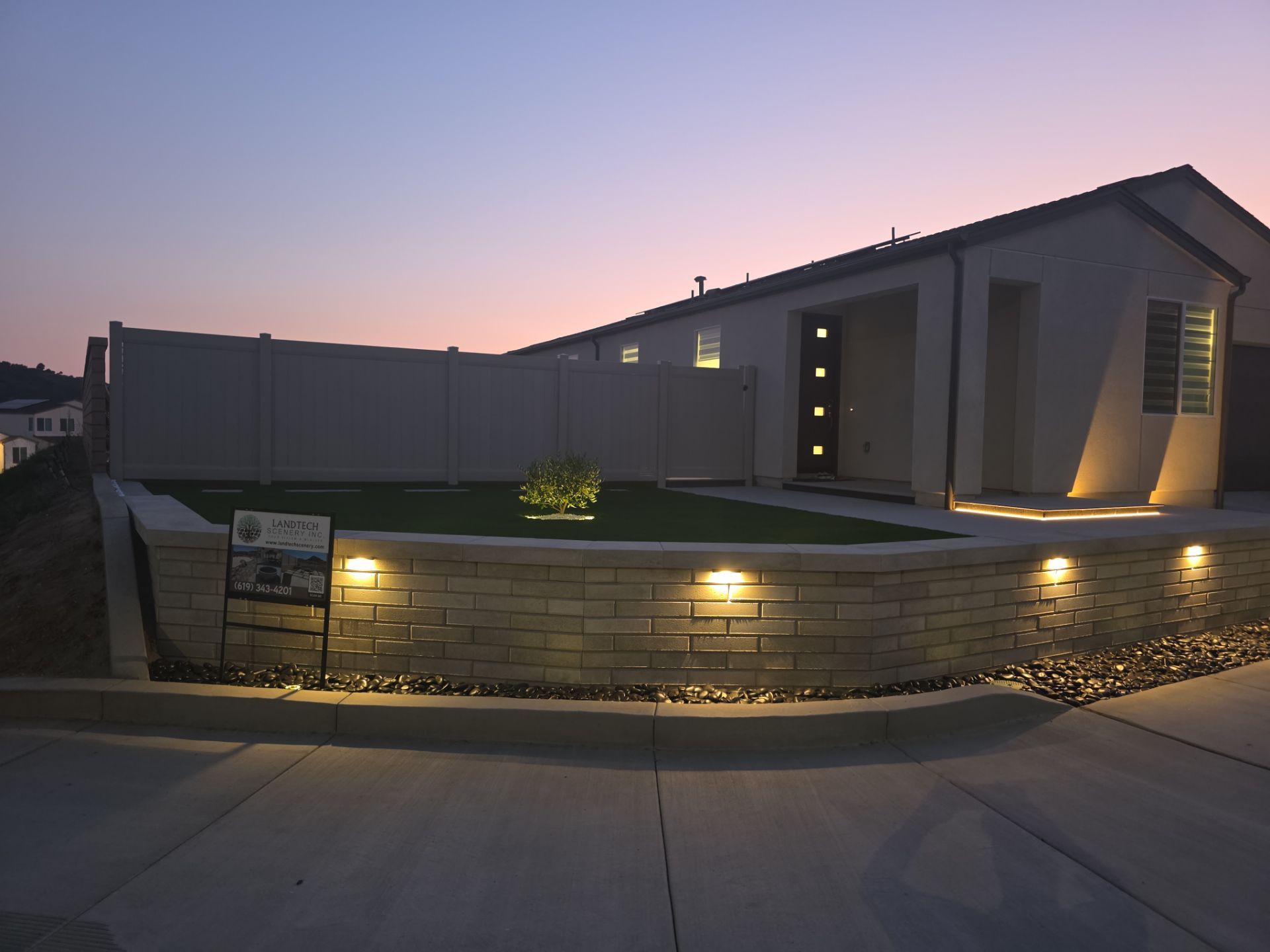 Exterior of a modern home at dusk with illuminated stone wall and lawn.