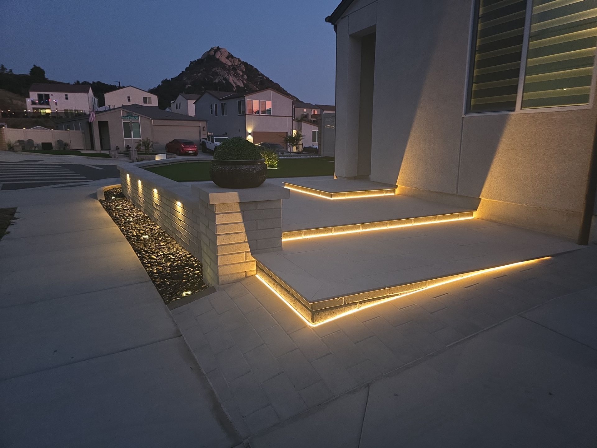 Lit exterior steps and low wall with embedded lights at dusk, residential neighborhood in background.
