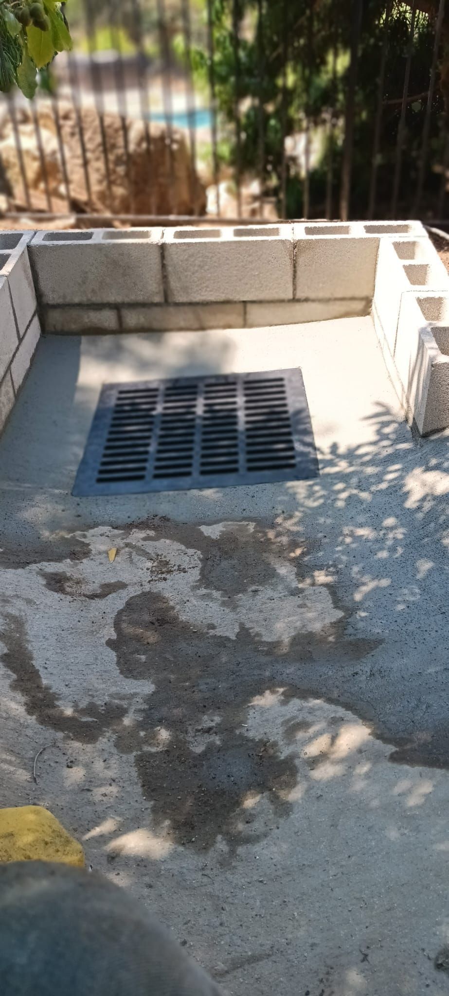 Concrete drainage grate set into a sidewalk beside a low cinderblock wall.