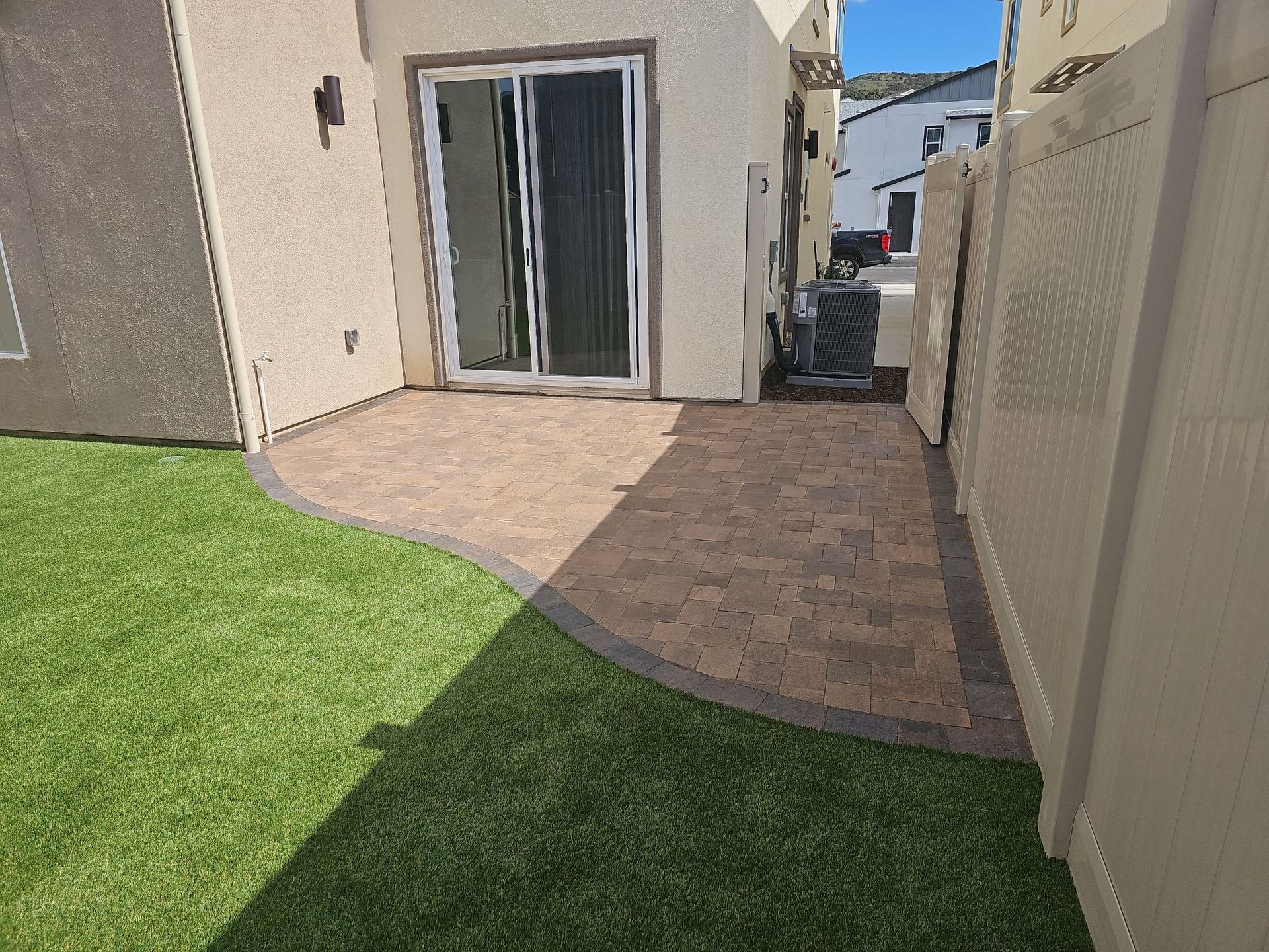 Patio with brick pavers, artificial grass, and sliding glass door. A beige fence is on the right.