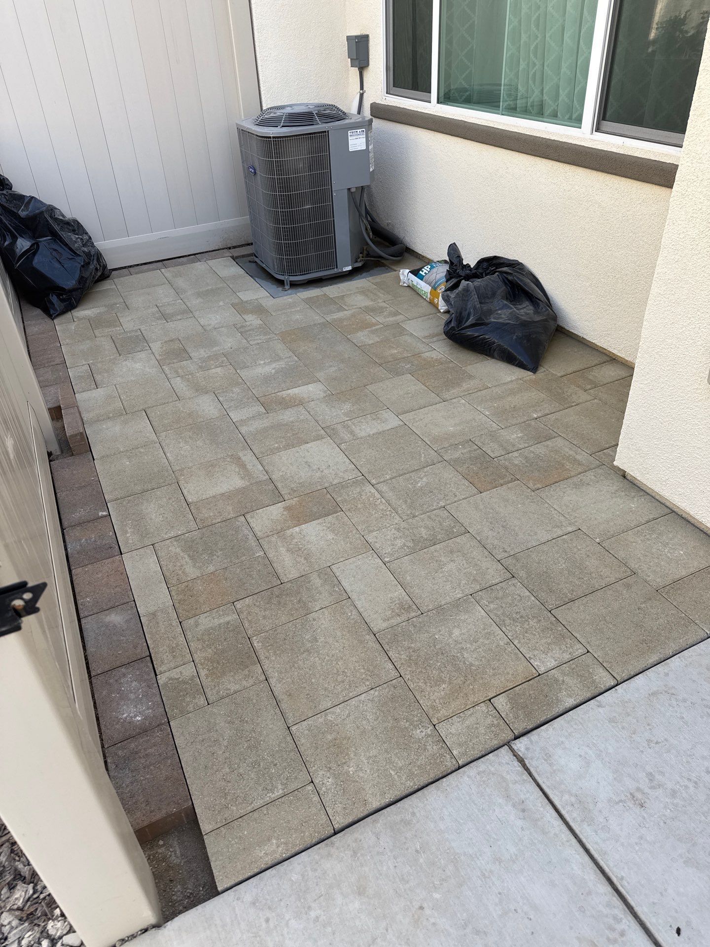 Patio paved with gray and brown bricks, with an air conditioning unit and trash bag in the corner.