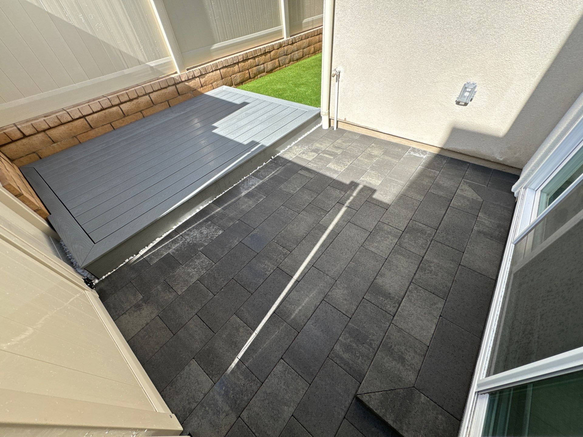 Patio with gray asphalt shingles and a gray elevated platform. Next to white house and fence.
