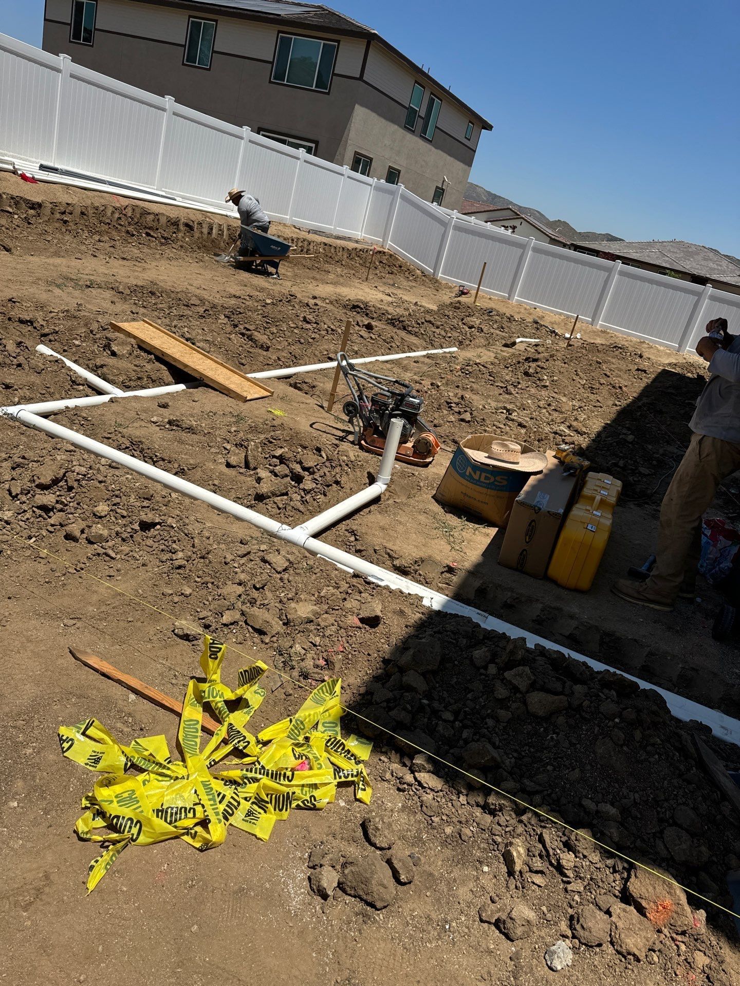 Construction site with white PVC pipes laid in dirt, white fence, house in background, bright sunny day.