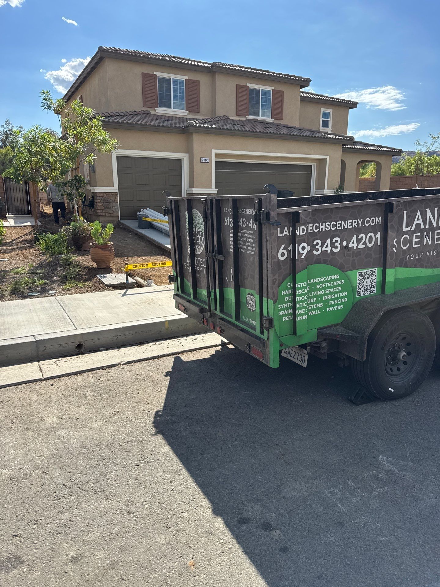 Green utility trailer parked in front of a two-story house on a sunny suburban street.