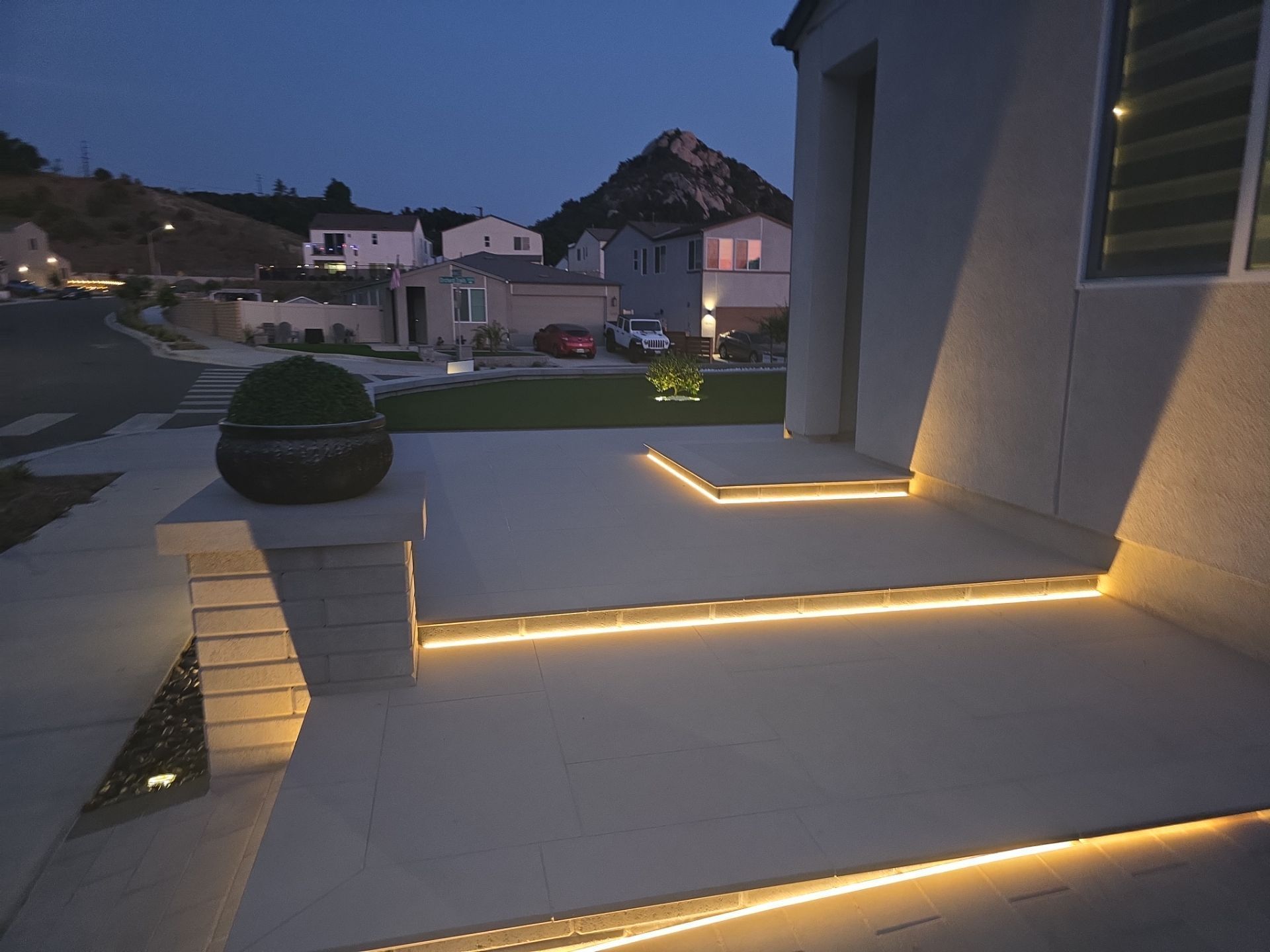 Lit concrete steps and porch at dusk. Yellow lights illuminate edges; houses and hills in background.