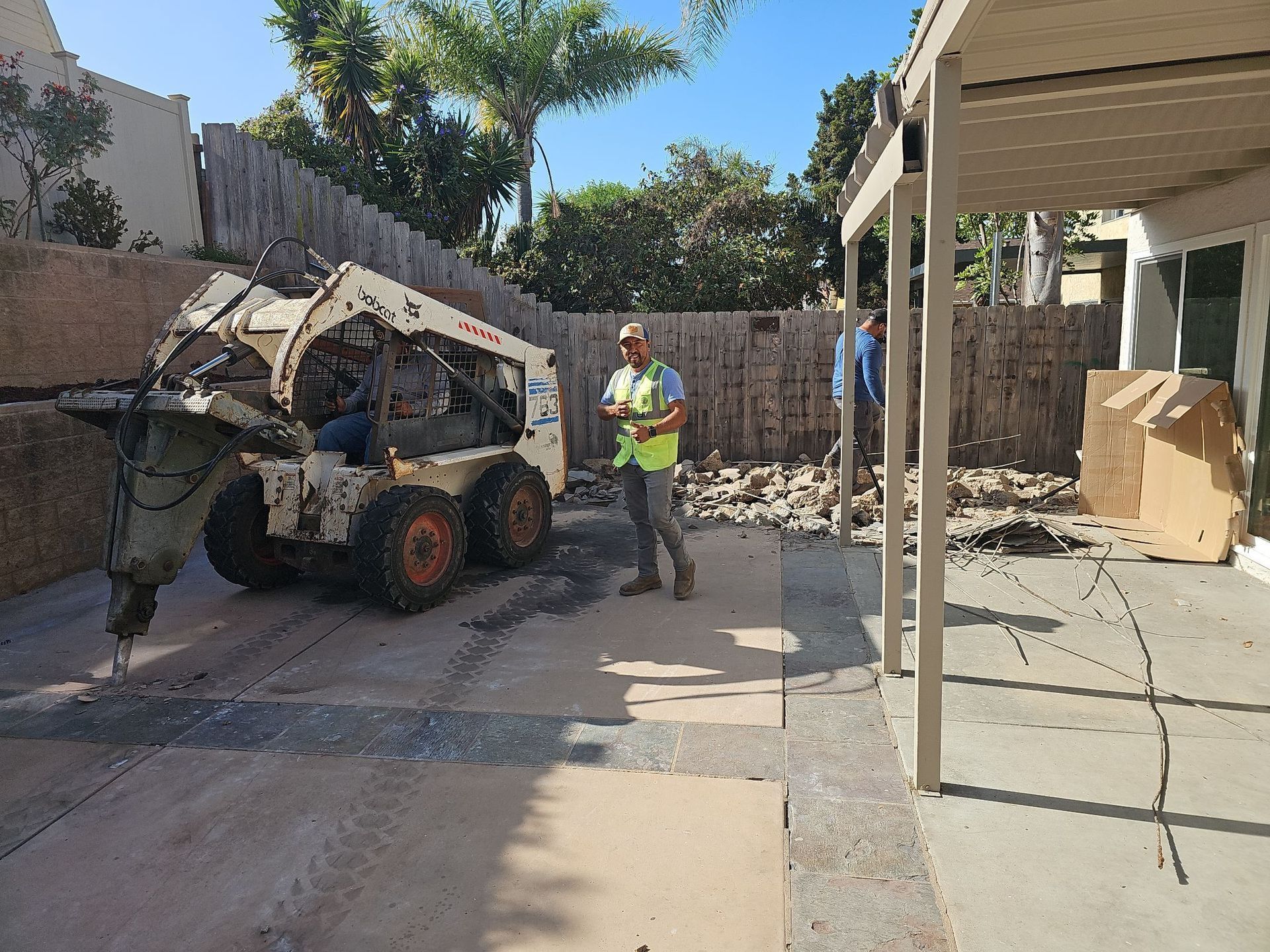 A Bobcat machine operating on a patio, two workers observe under the shade.