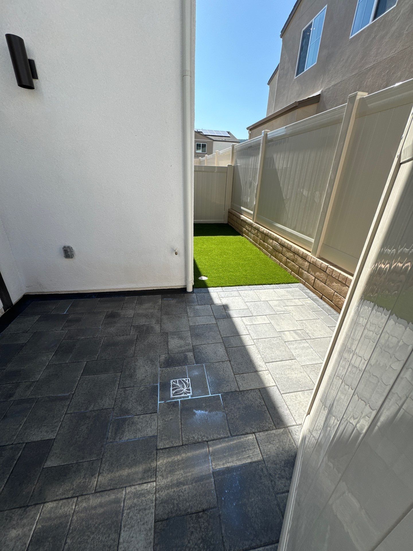 Patio with dark gray pavers, leading to a small grass area, with a white wall and tan fence.
