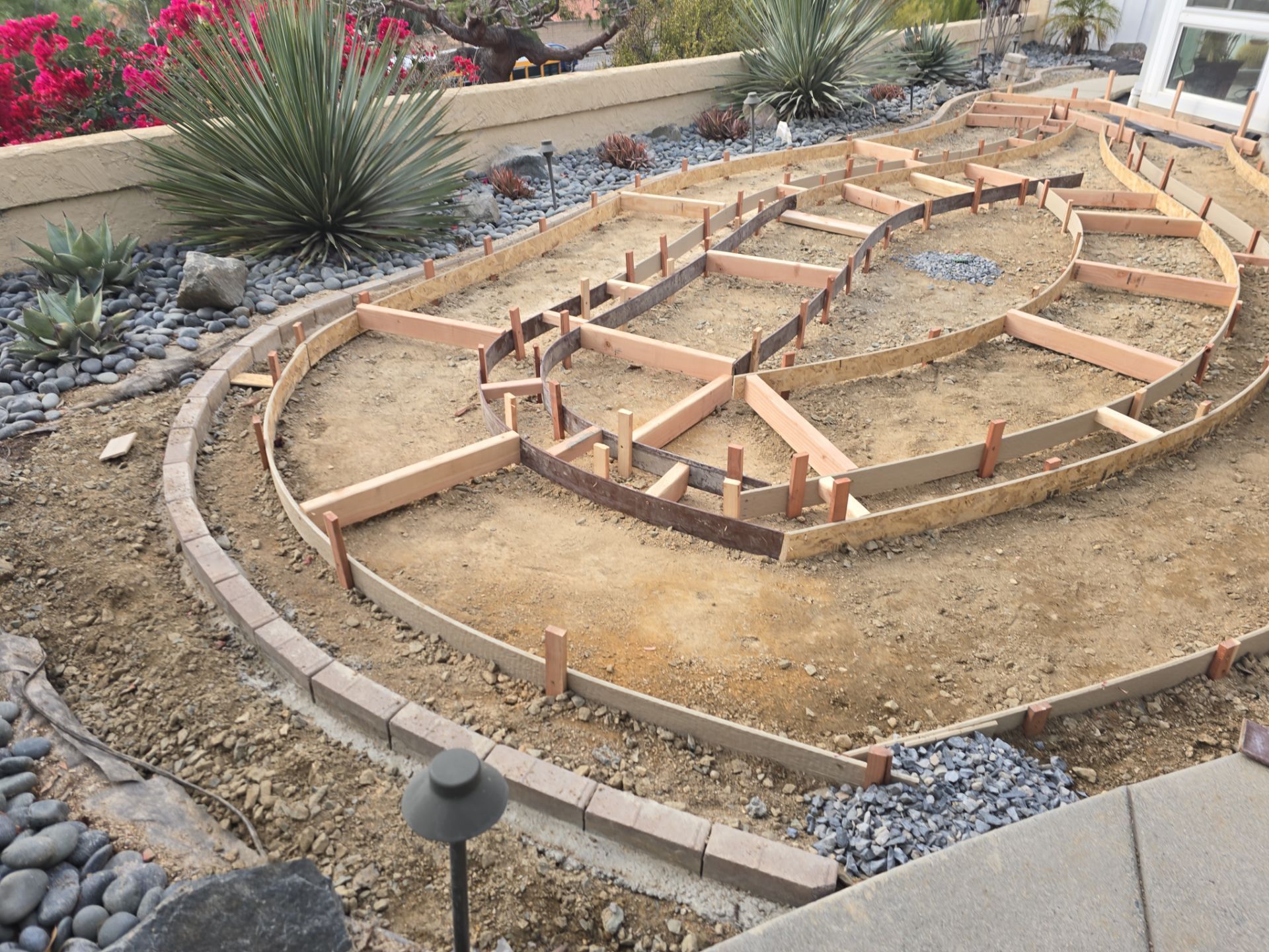Wooden forms outlining a tiered garden bed in a dirt area, with a stone border and existing landscaping.