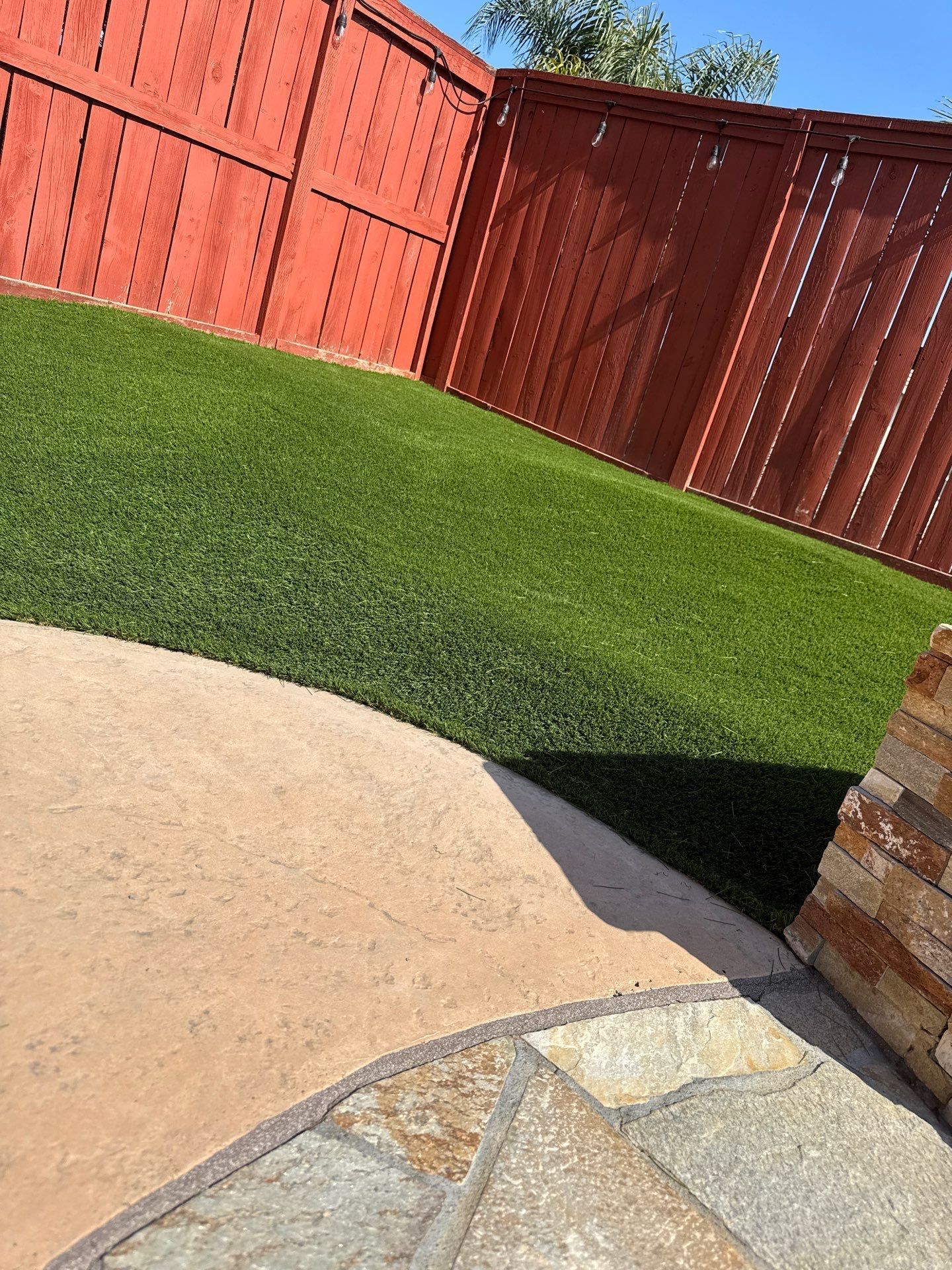 Lawn with red fence, bordering a patio made of flagstone and concrete, sunny day.
