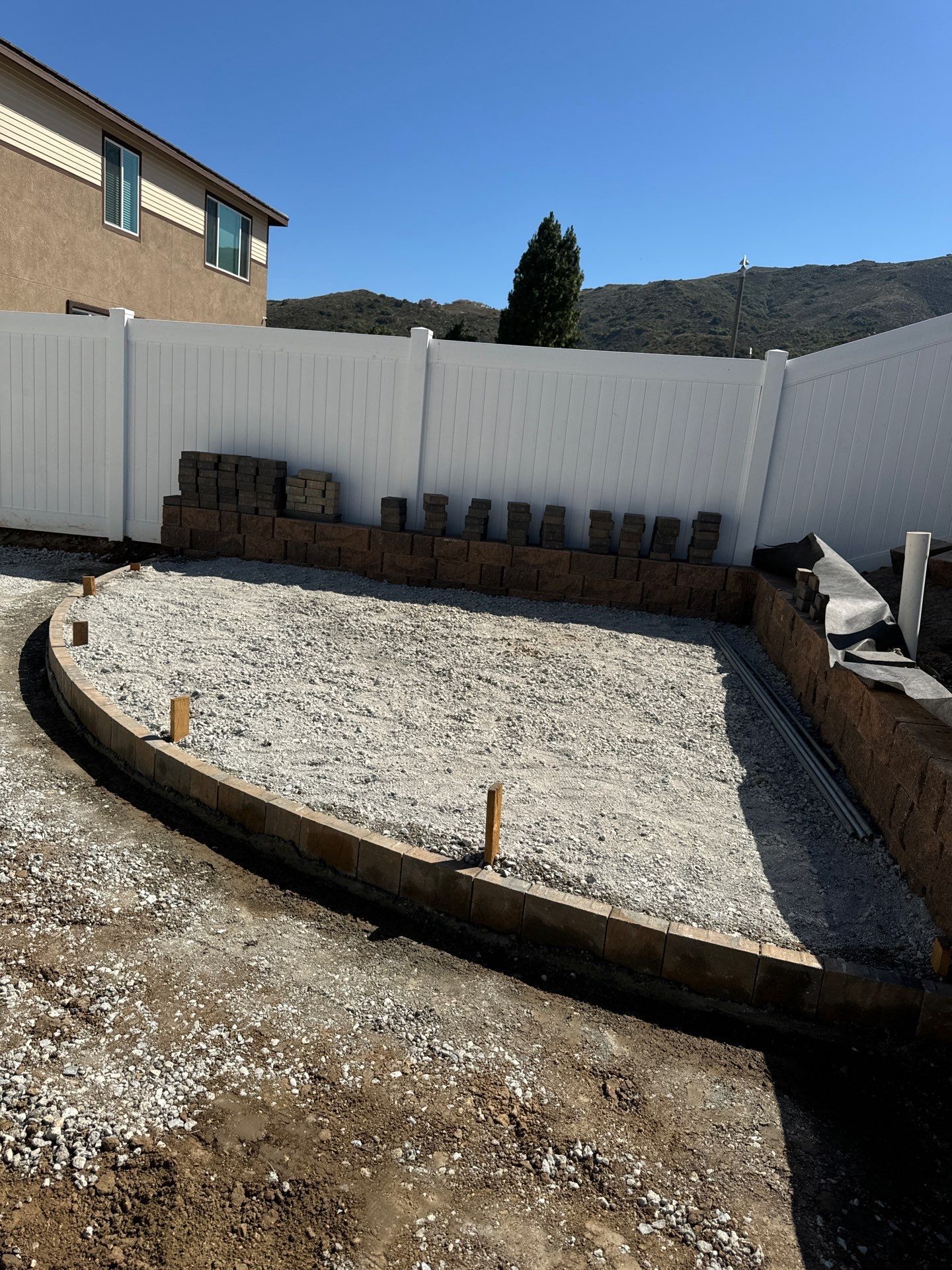 Backyard construction: gravel base, curved wooden border, stacked bricks, white fence, clear sky.