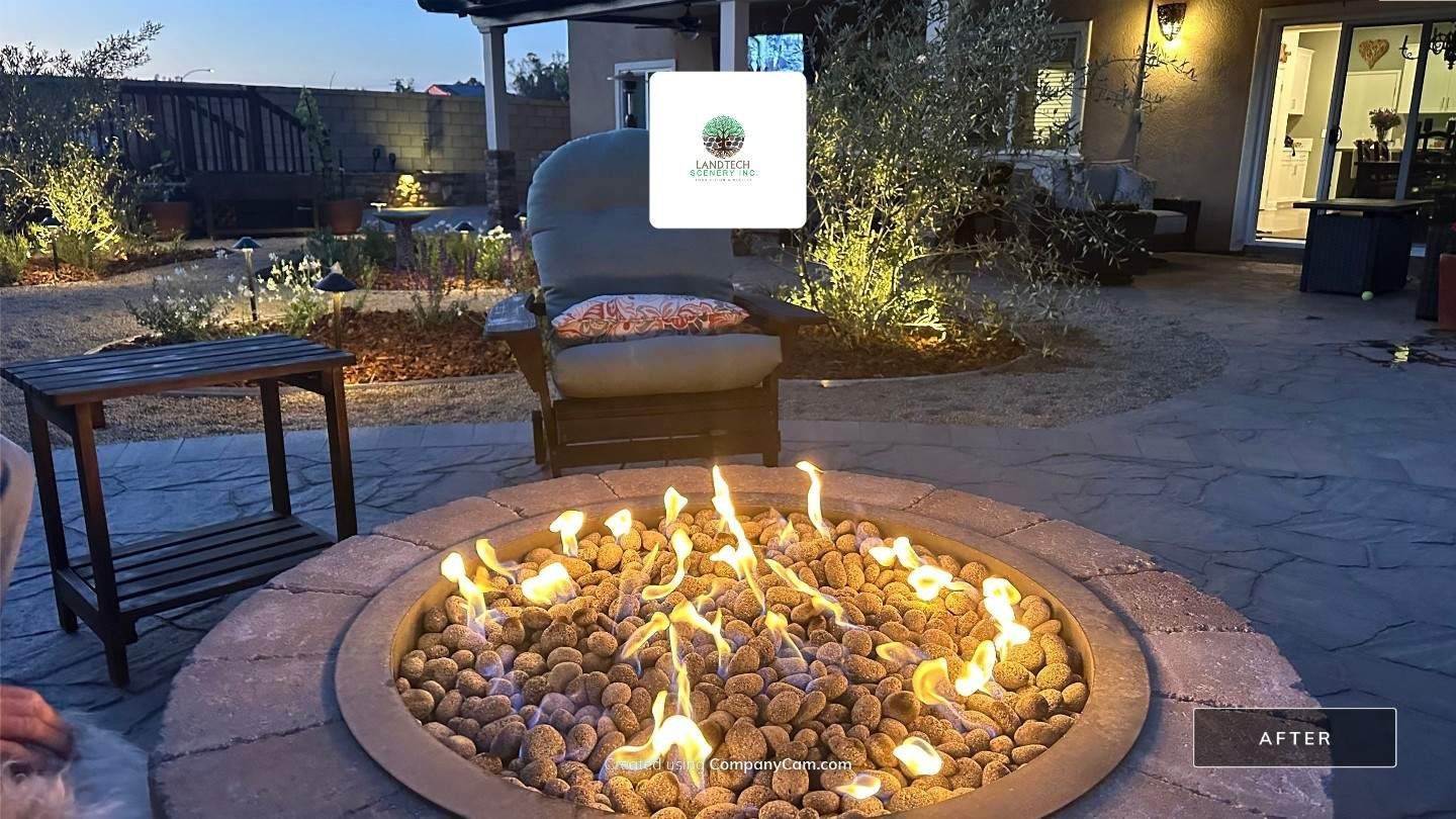 Backyard patio with a circular fire pit burning at dusk, surrounded by chairs and a small table.