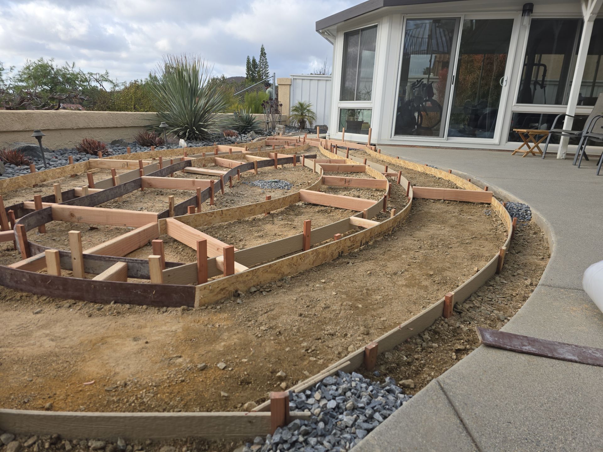 Construction of a curved garden bed with wooden frames, on dirt, next to a patio and house.