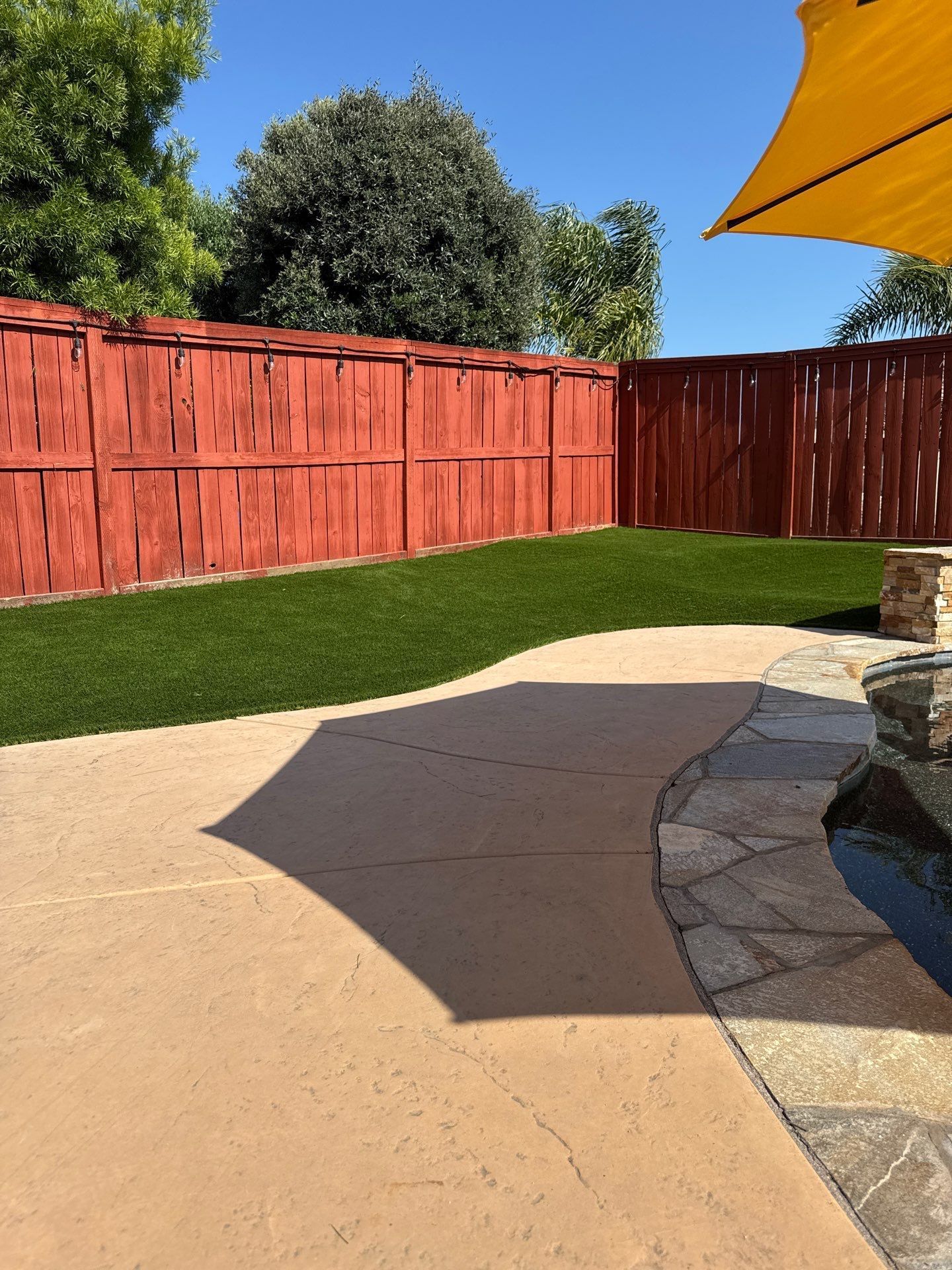 Backyard with red fence, green lawn, paved patio, yellow umbrella, and a water feature.
