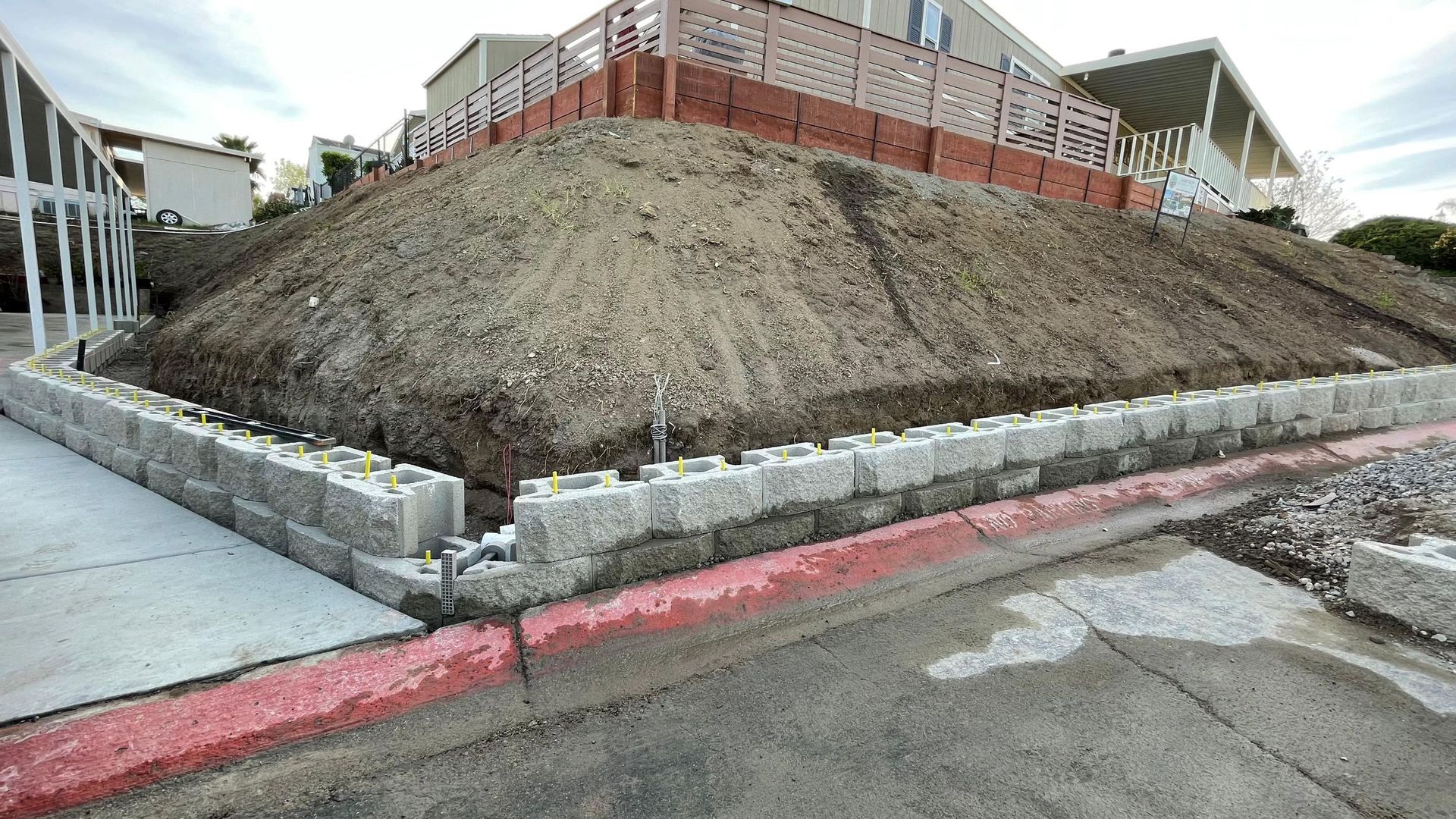 Retaining wall made of gray blocks supporting a mound of dirt next to a red-lined road.