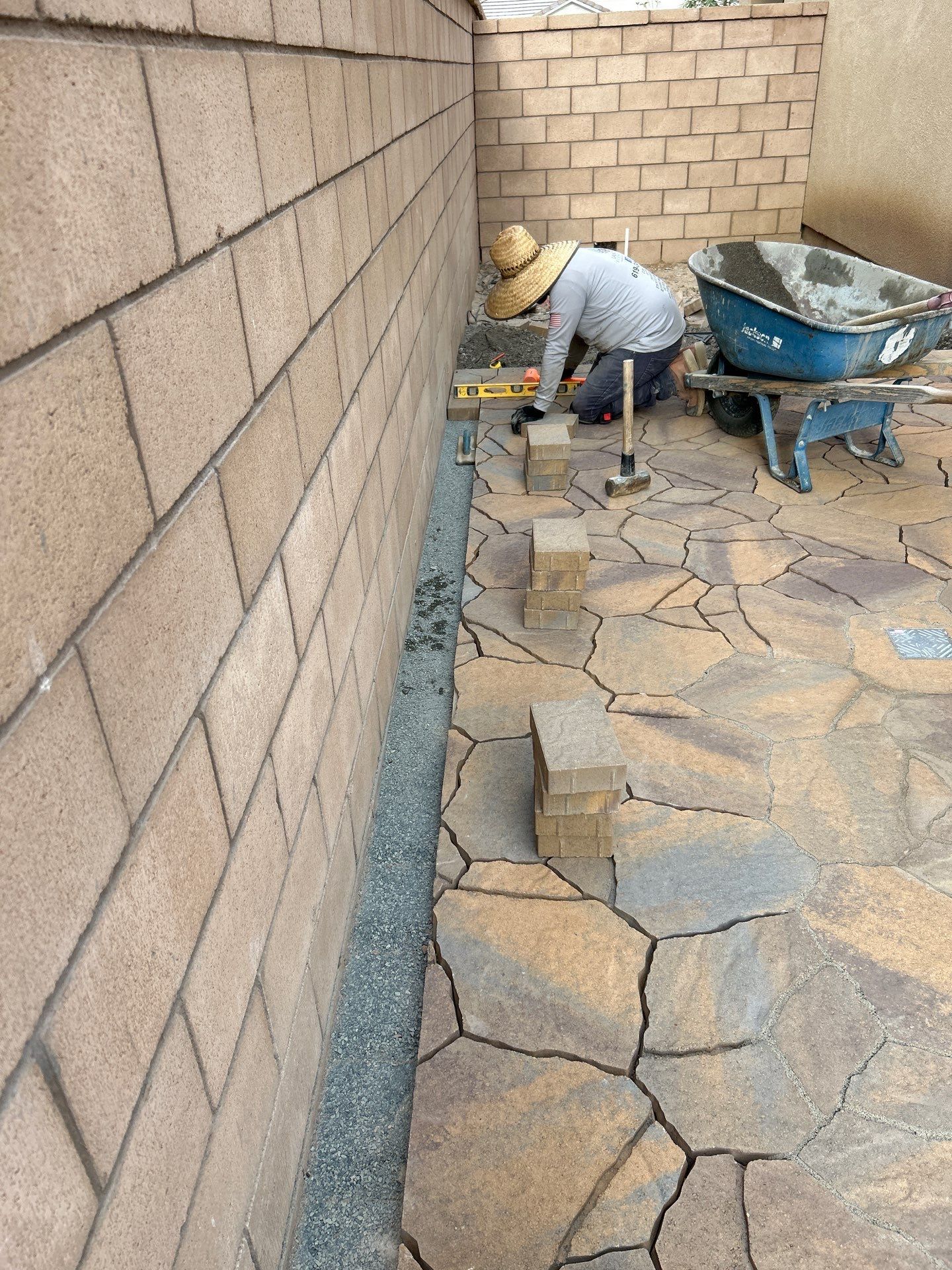 Worker laying stone pavers beside a brick wall, with a wheelbarrow in a narrow outdoor courtyard.