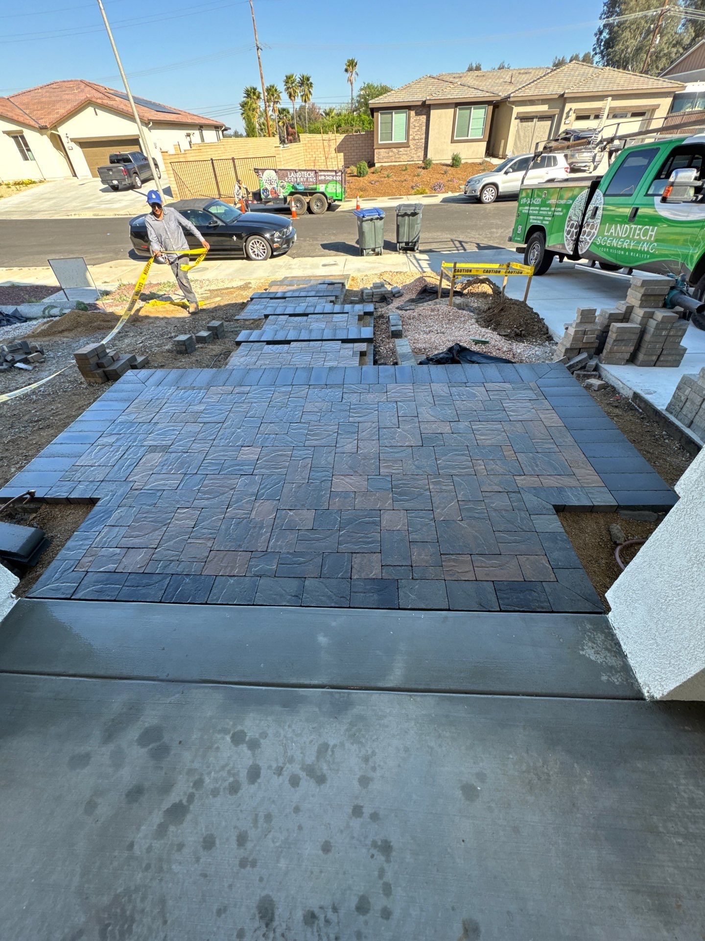 Paver installation in progress; driveway with brick pavers, worker using level. Green truck in background. Sunny day.