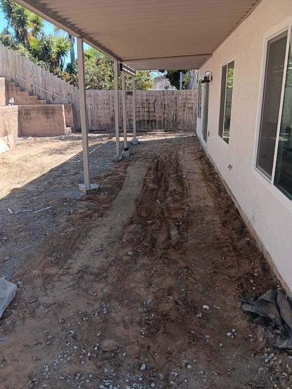 Patio with a metal roof, dirt ground, and a white house with windows. Wooden fence in the background.