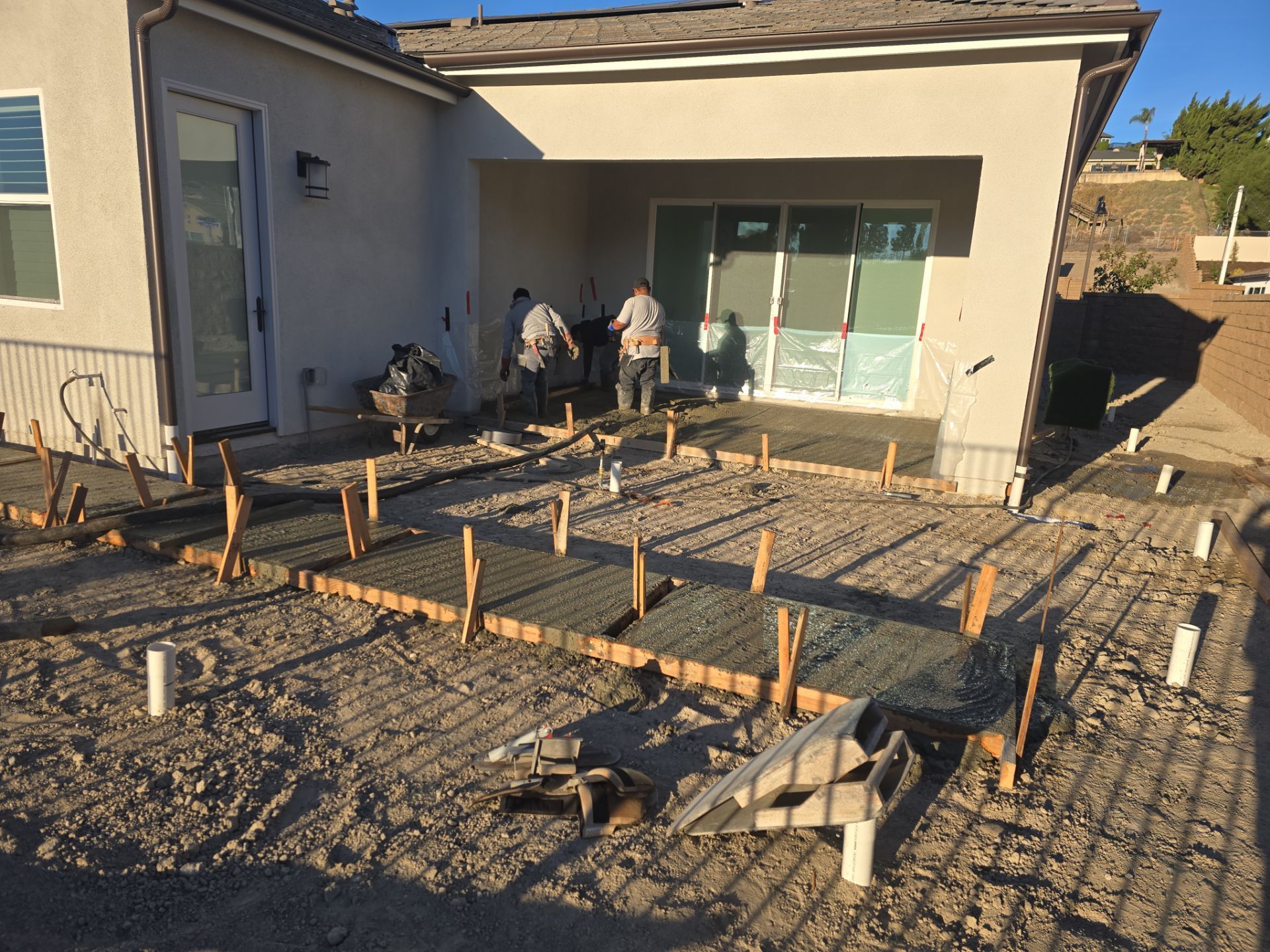 Workers pouring concrete for a patio. Wooden forms, rebar, and plumbing visible. Exterior of a house in the background.