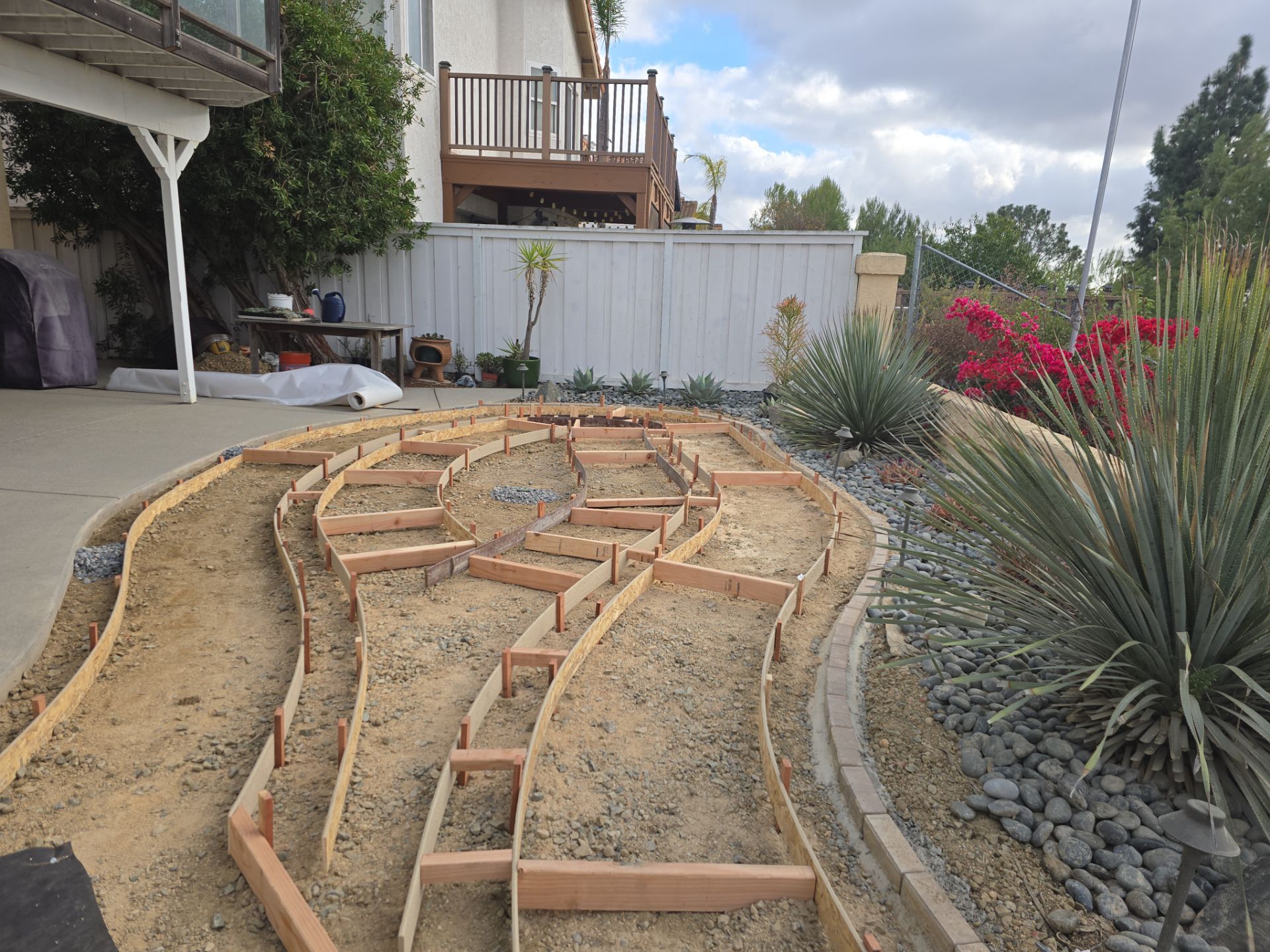 Wooden forms delineate tiered garden beds filled with dirt, alongside a gravel path and mature landscaping.