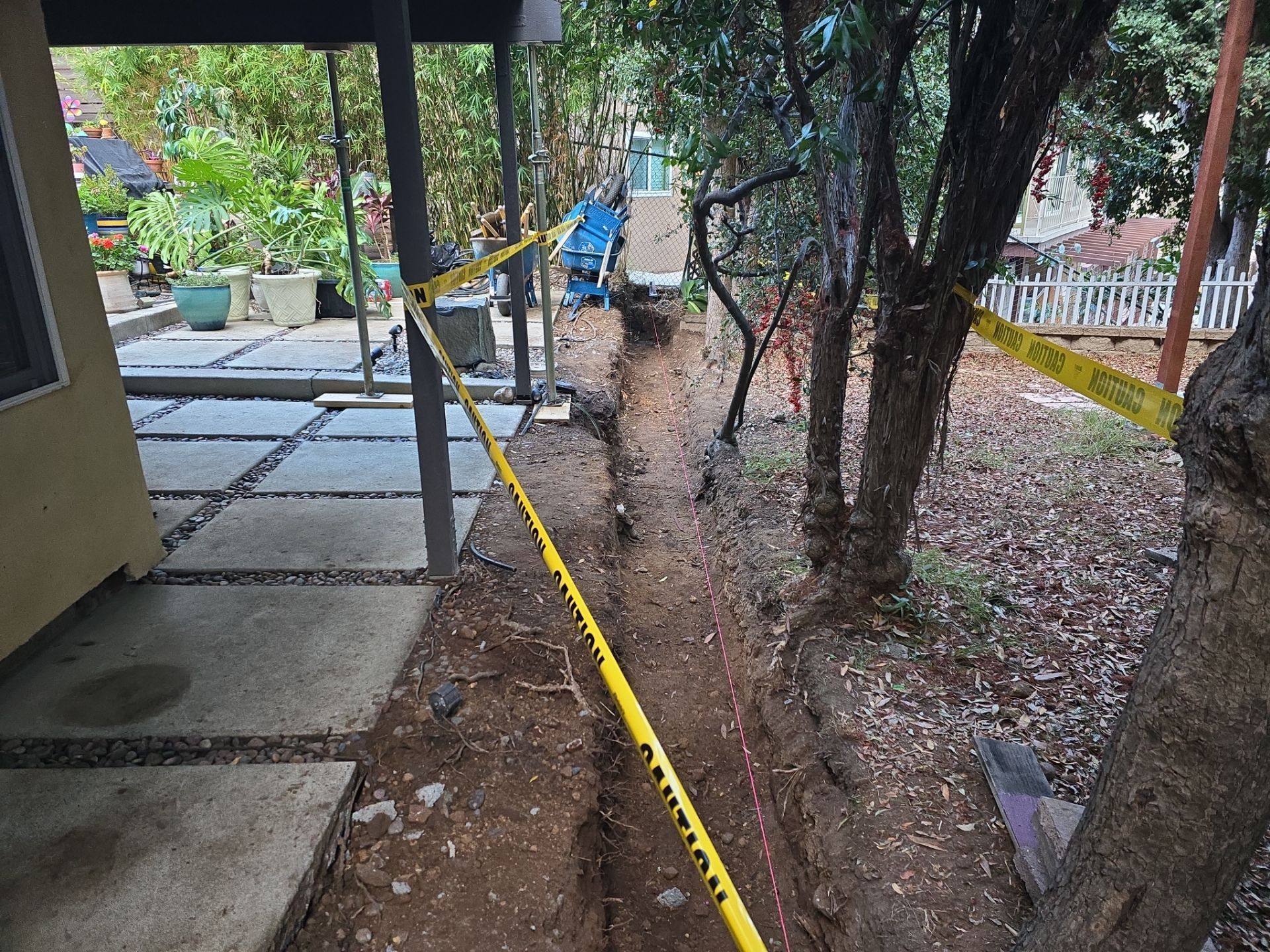 A trench with yellow caution tape, between a house and trees, likely for construction.