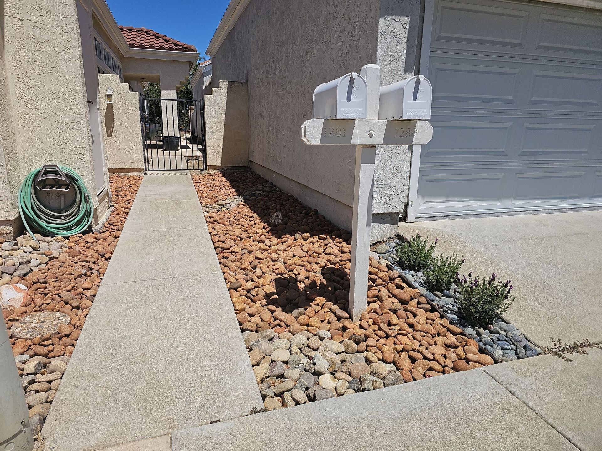 Concrete pathway to a front gate with a mailbox. Landscaping includes rocks and greenery.