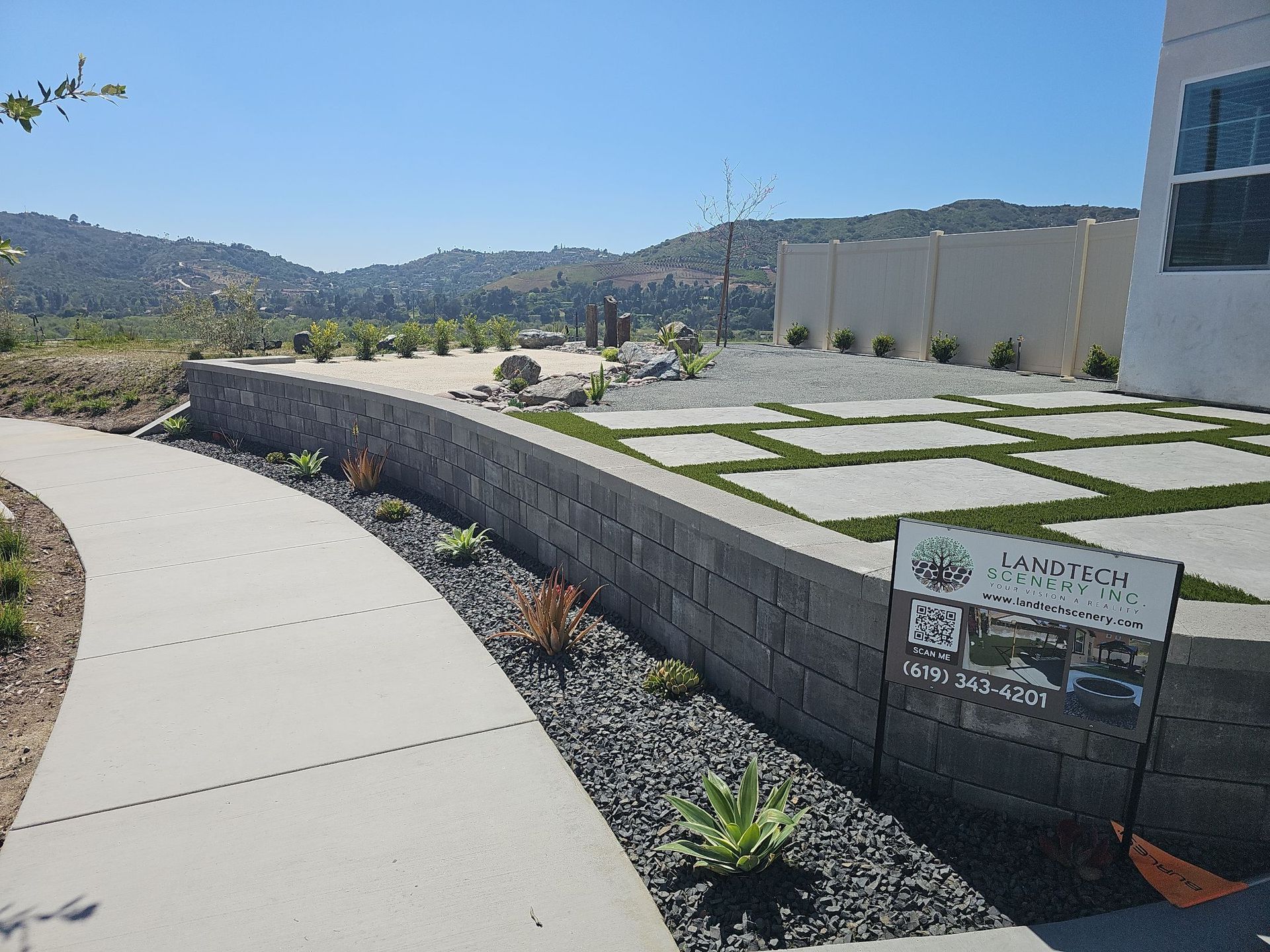 Curved concrete walkway next to a gray stone retaining wall with landscaping and a modern house.