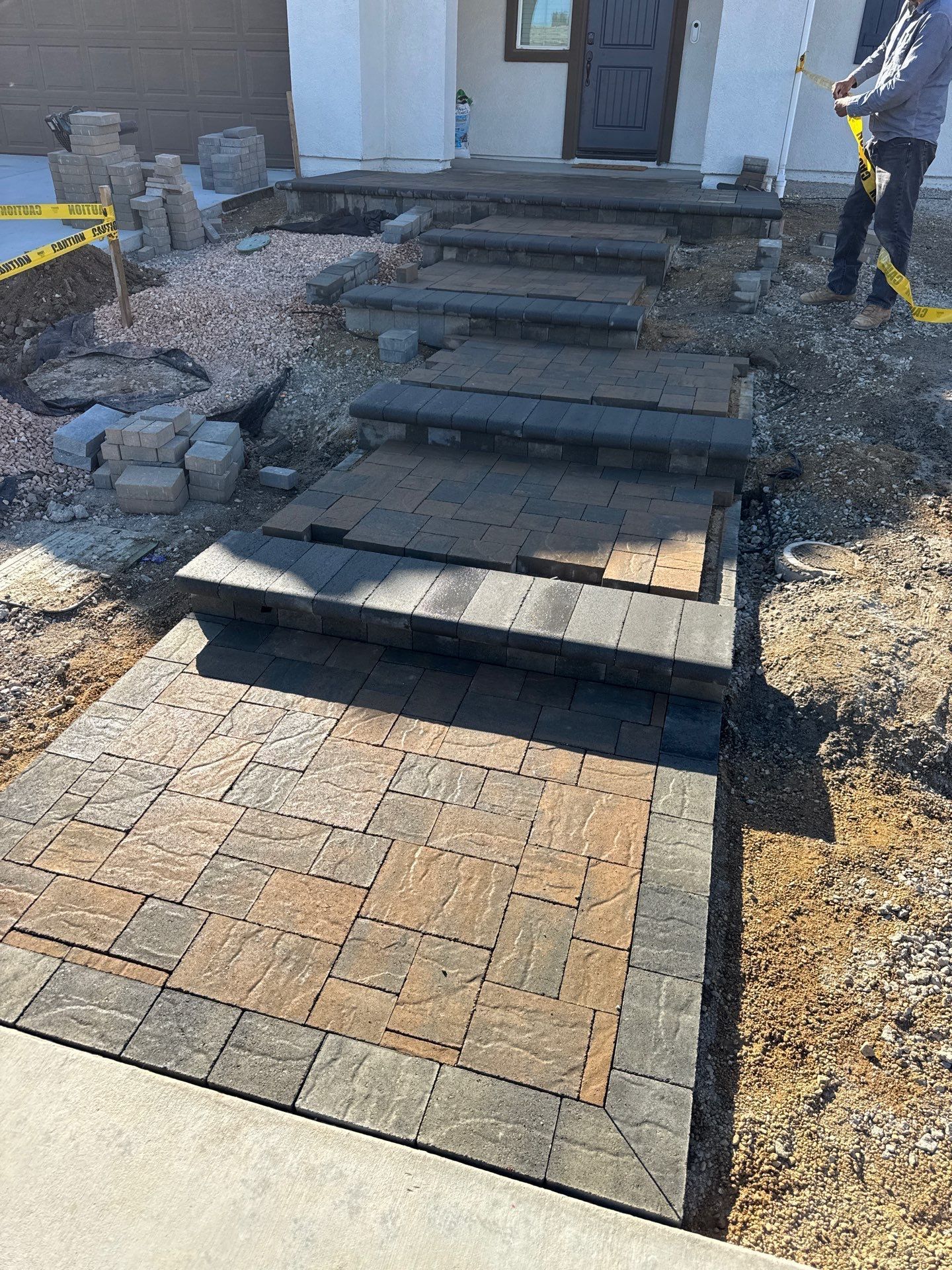 Brick steps and walkway being constructed in front of a house; brown and gray pavers, work in progress.