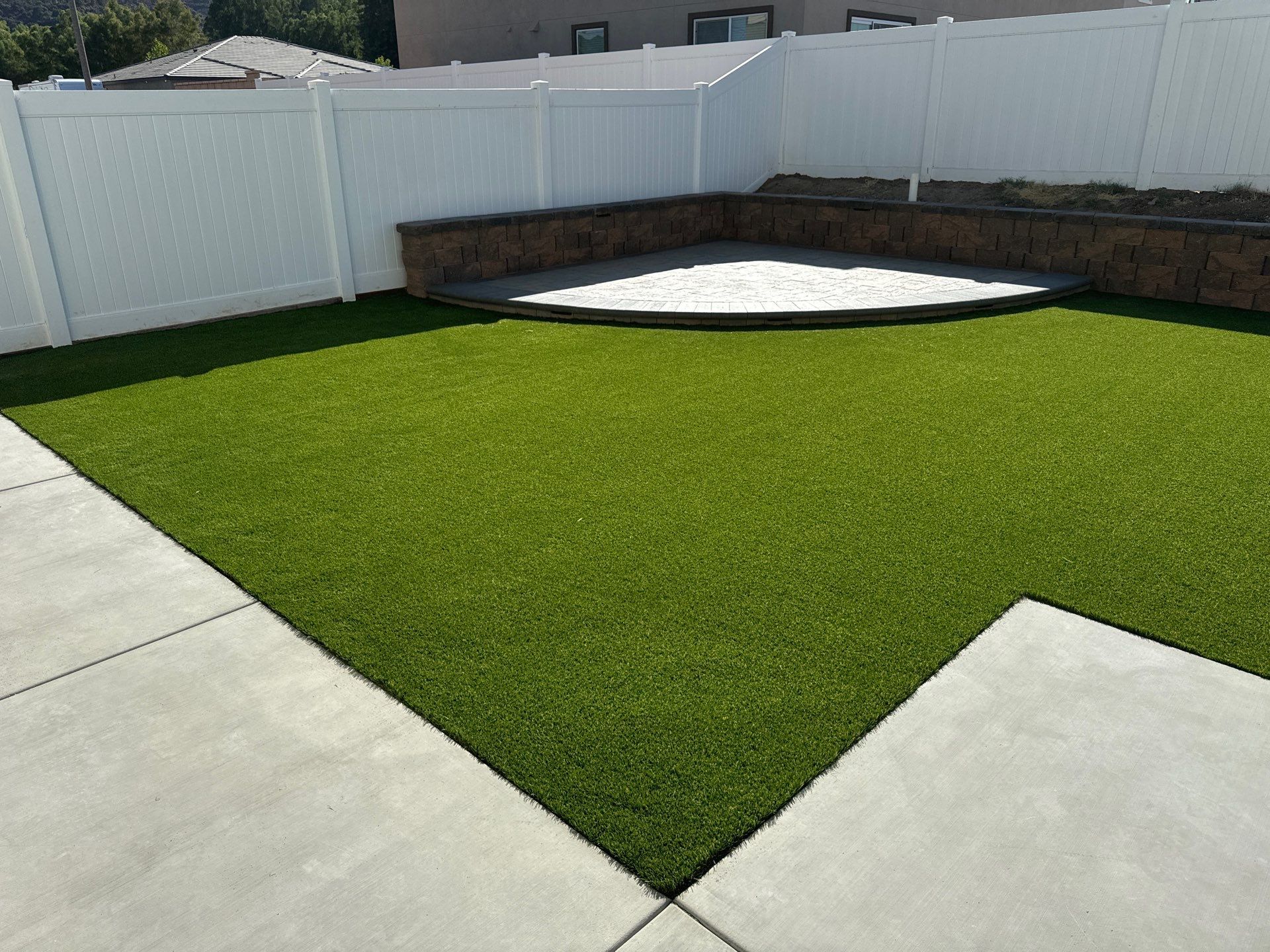 A backyard with a rectangular concrete patio, green artificial turf, and a raised brick-lined area with white fencing.