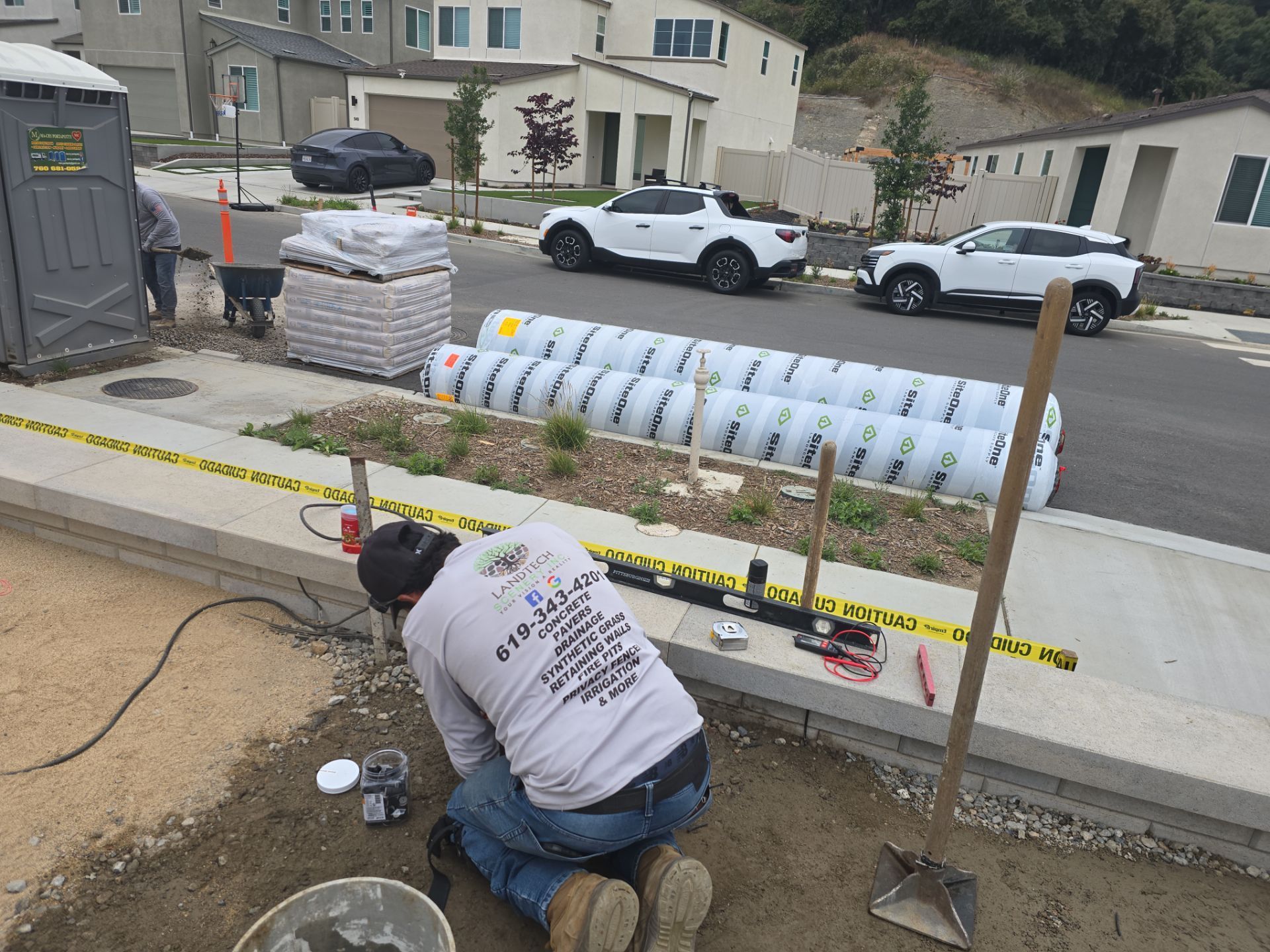 Worker kneeling on a sidewalk beside construction materials and tools near a residential street.