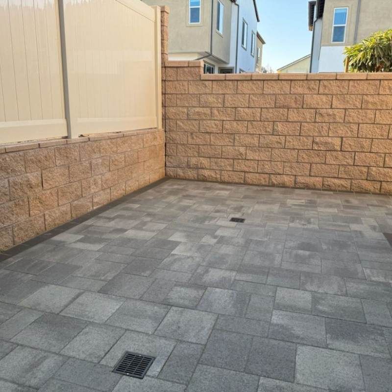 Enclosed patio with gray pavers, brick walls, and a white fence. Drainage grates are visible.