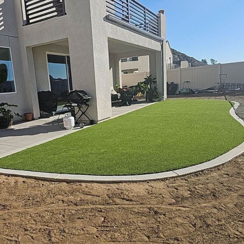 A backyard with a green artificial lawn bordering a concrete patio near a house with a balcony.