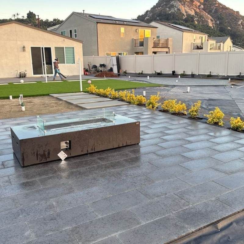 Patio with fire pit, pavers, artificial turf, and small shrubs. Person in backyard.