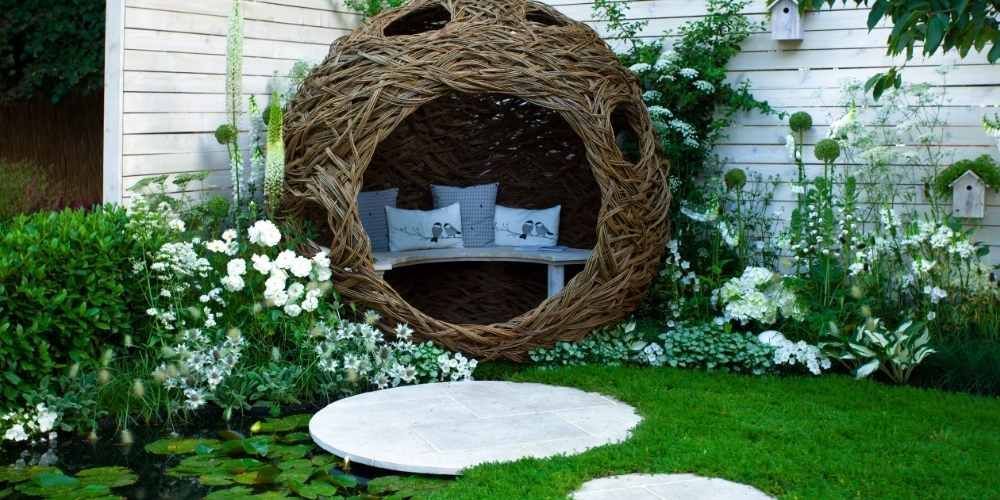Wicker nest-like outdoor seating with pillows, set in a garden with stepping stones and white flowers.