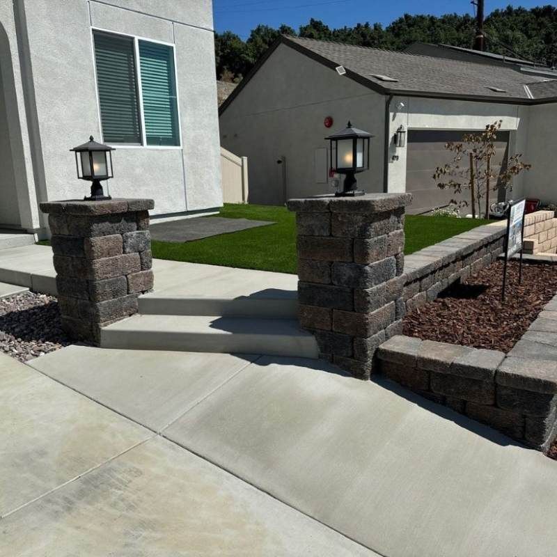 Stone pillars with lamps flank steps leading to a house with a concrete walkway.