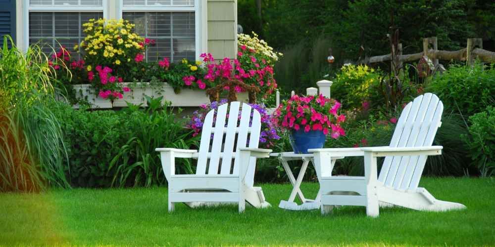 Two white Adirondack chairs and small table on green lawn, pink flowers, yellow flowers, and garden background.