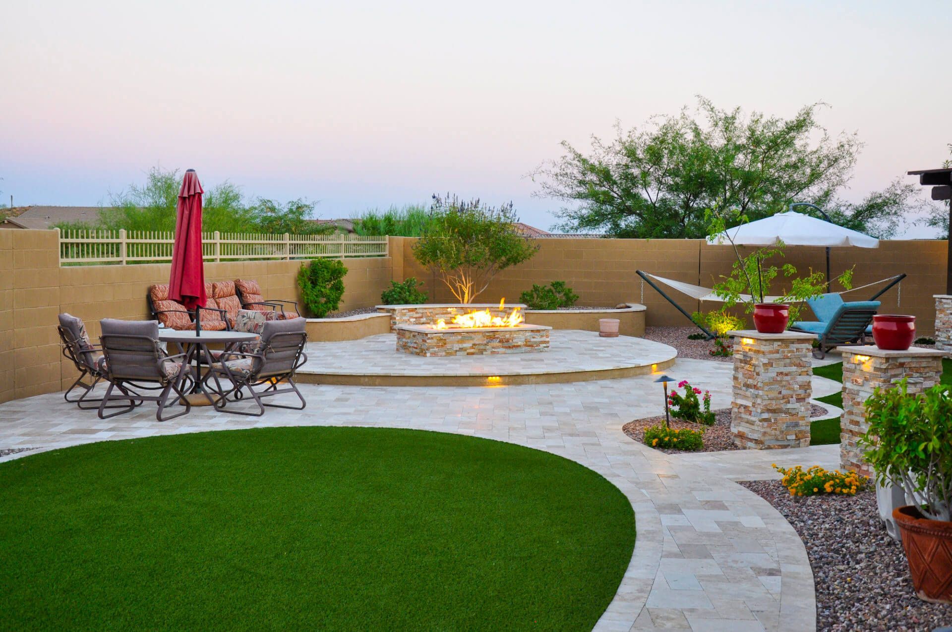 Backyard patio with fire pit, seating, hammock, and artificial lawn at sunset.