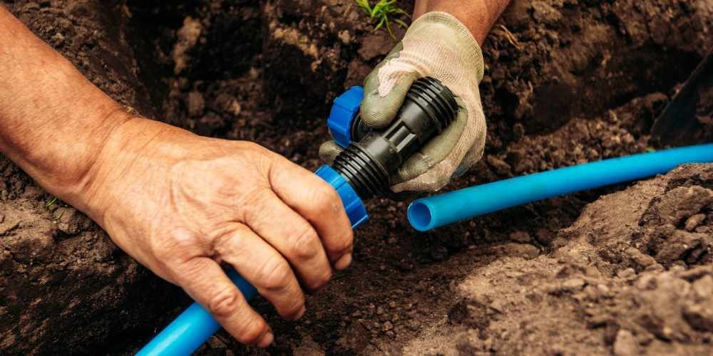 Hands connecting a blue irrigation hose to a sprinkler valve in soil outdoors